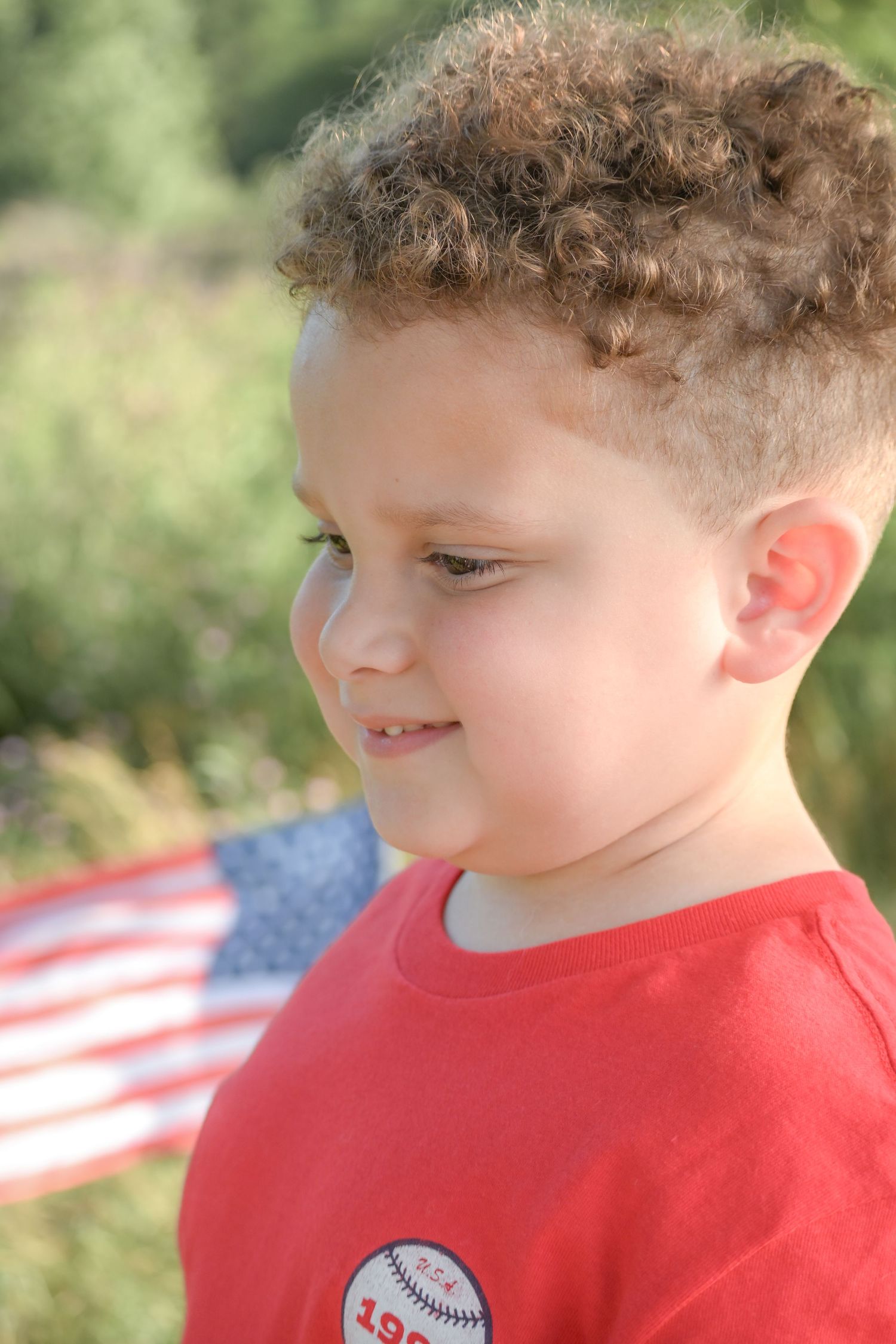 Curly-haired young boy in red baseball shirt looks down with American flag in background.