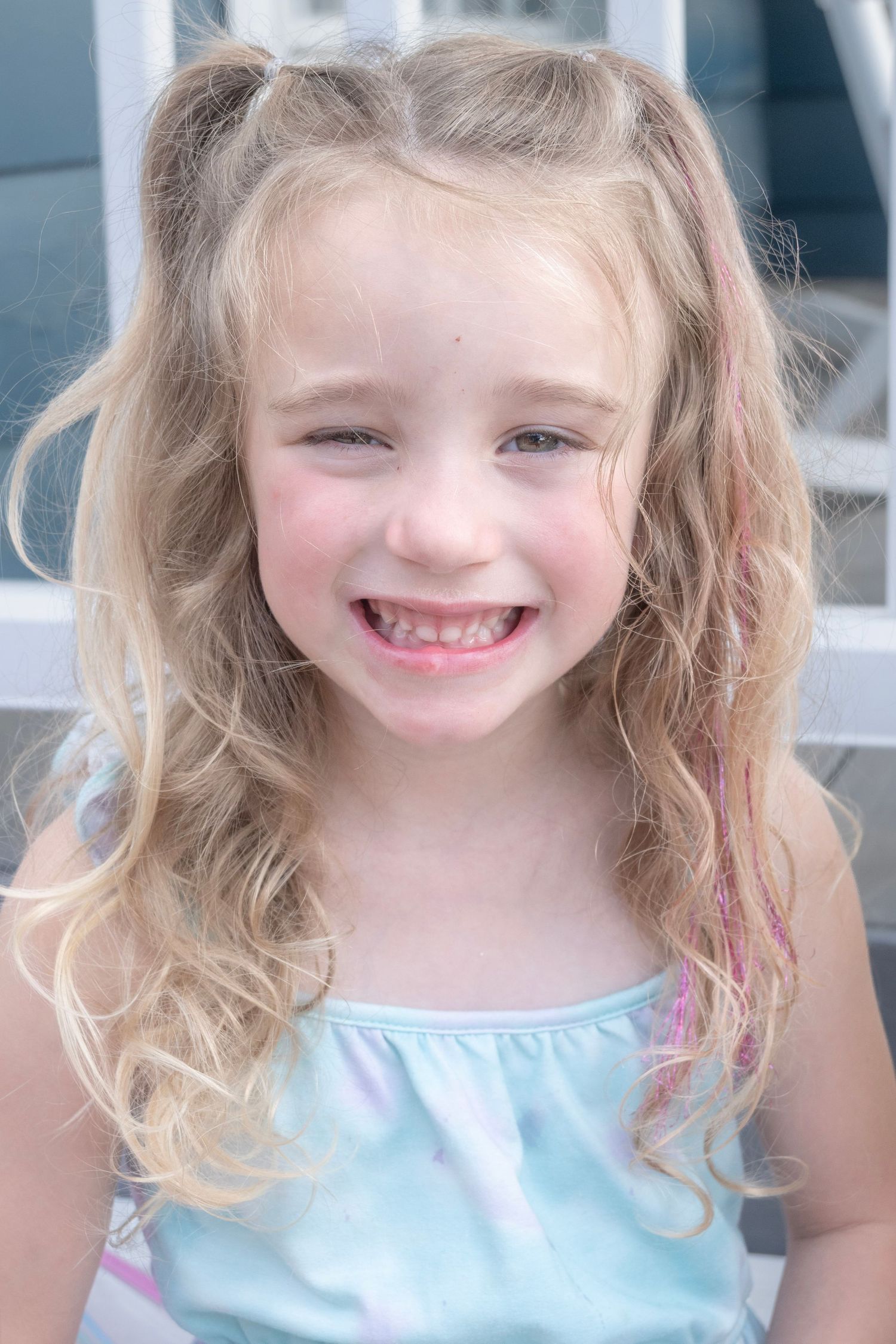 Smiling young girl with long blonde curly hair and pink highlights wearing a pastel blue top.