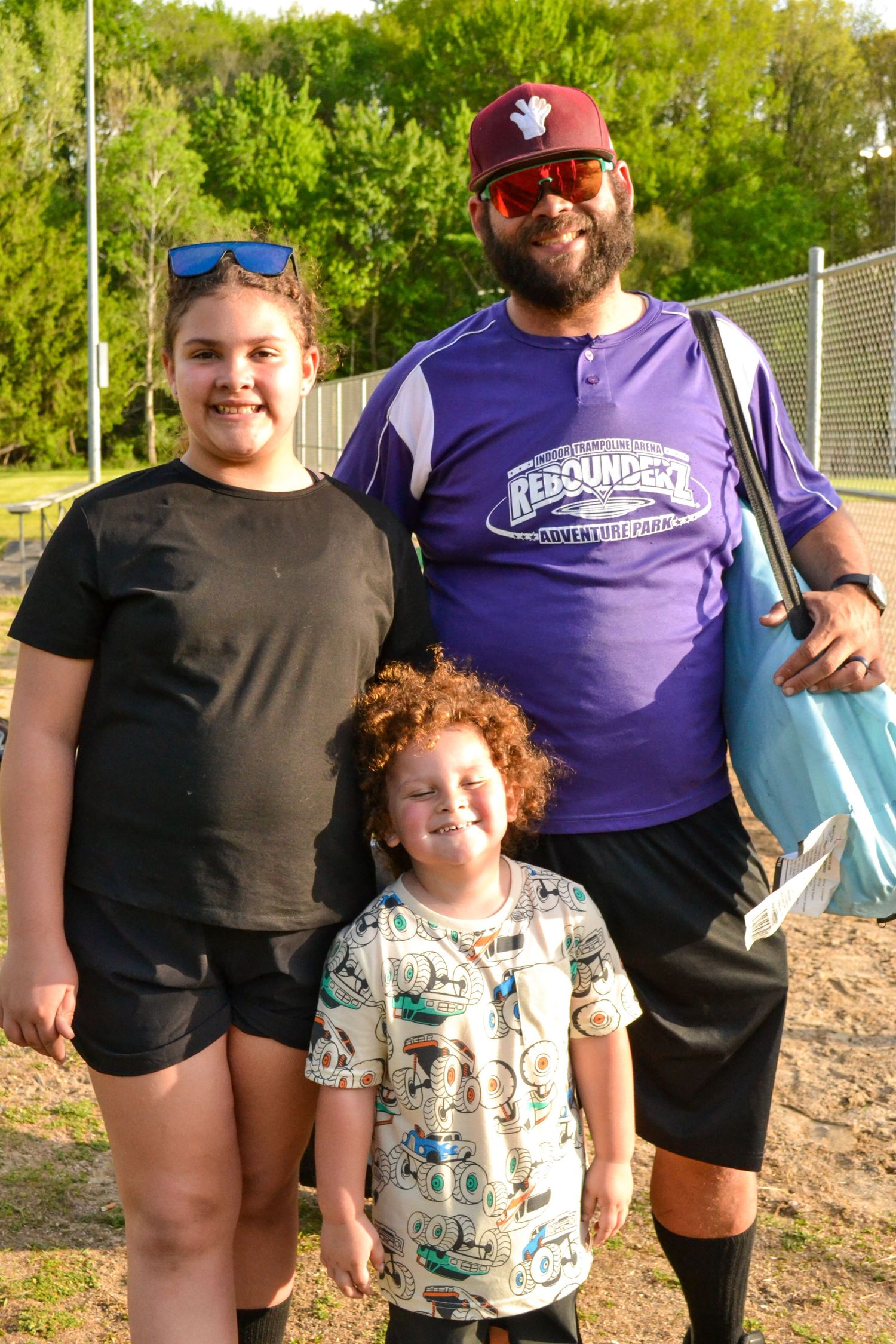 Man with beard and two young girls smiling together outdoors at a sports field.