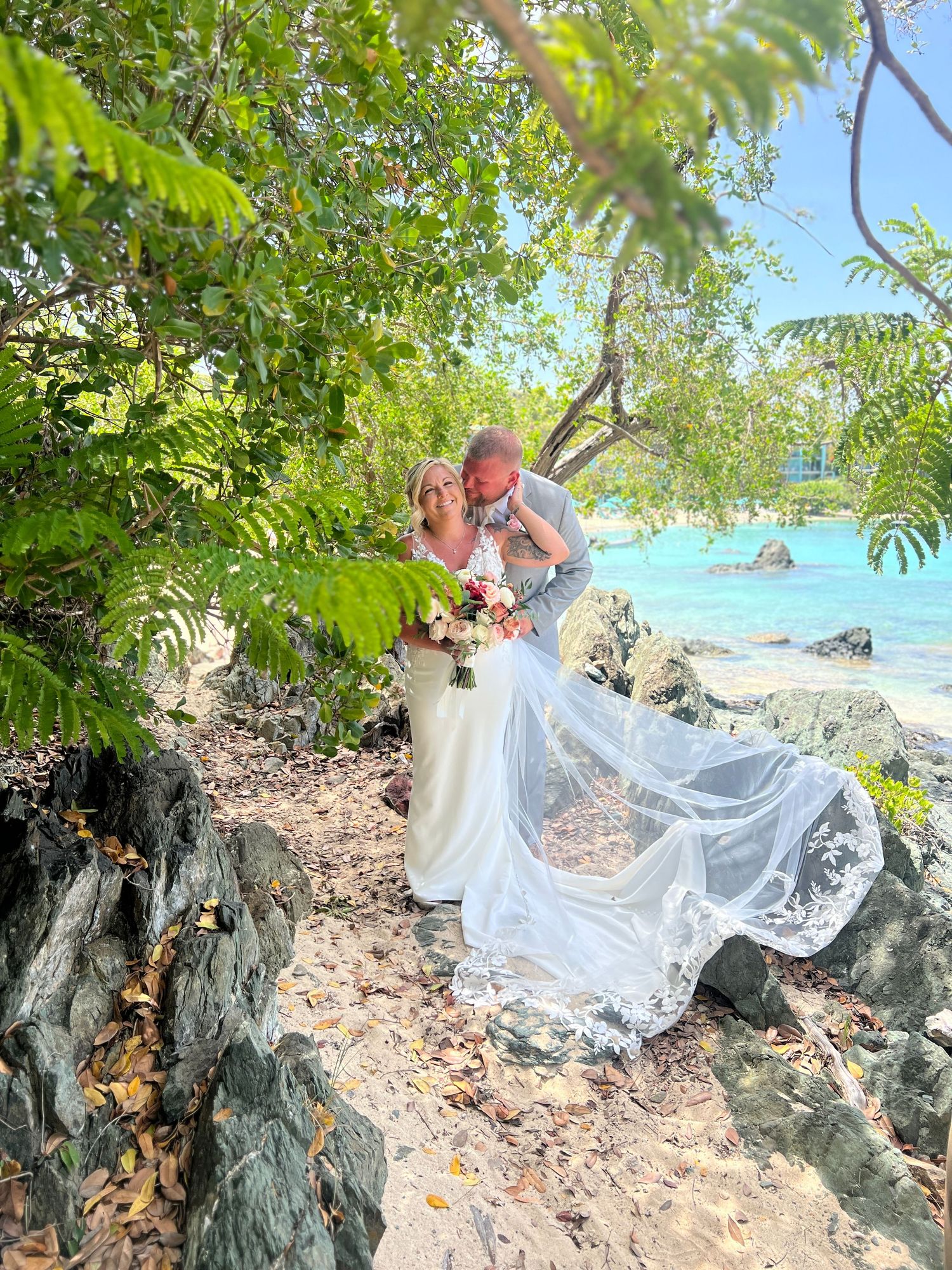 Bride and groom sharing a romantic dip kiss among lush tropical trees near a stunning turquoise beach.