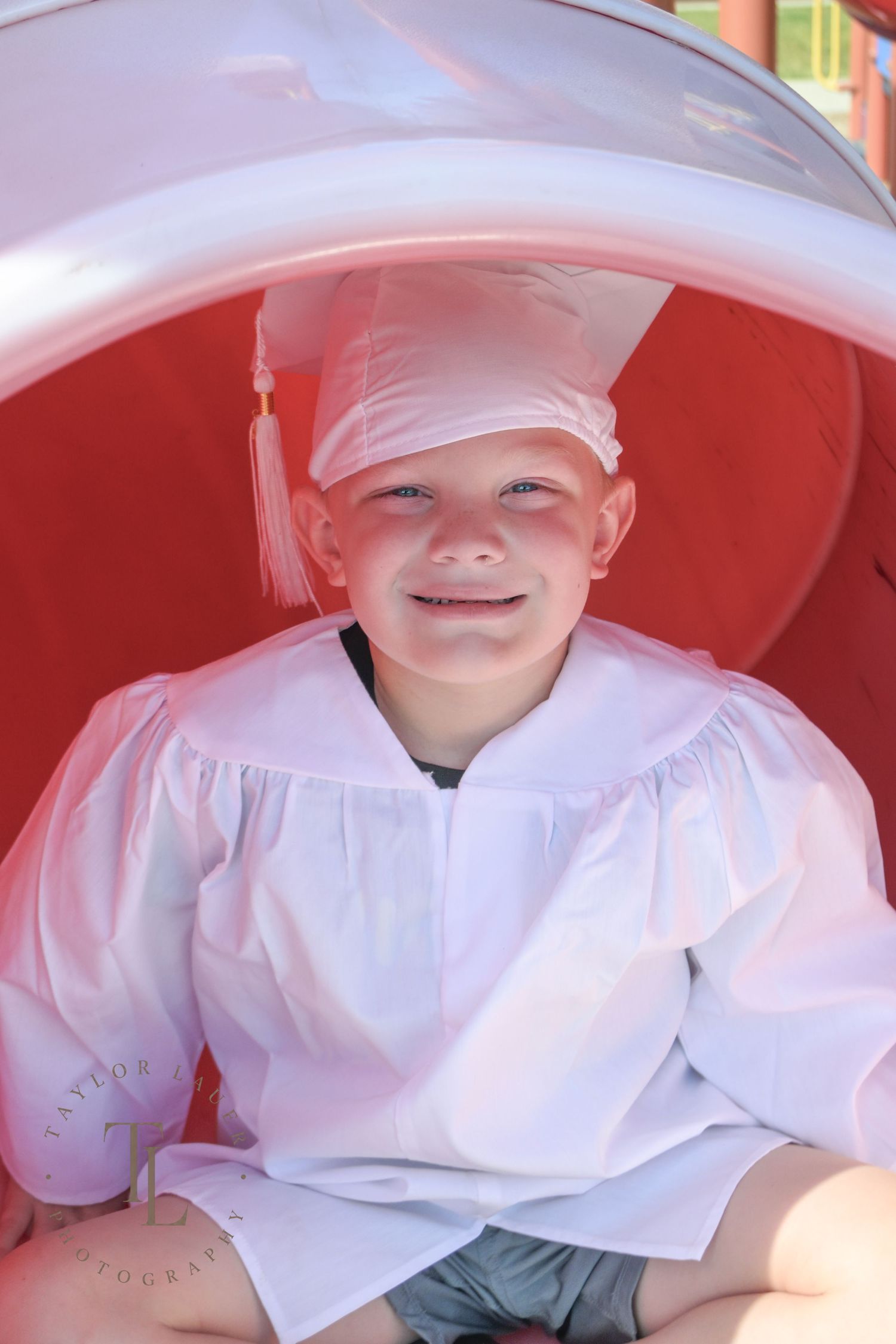 Young boy smiling in white graduation cap and gown sitting inside a red playground tunnel.