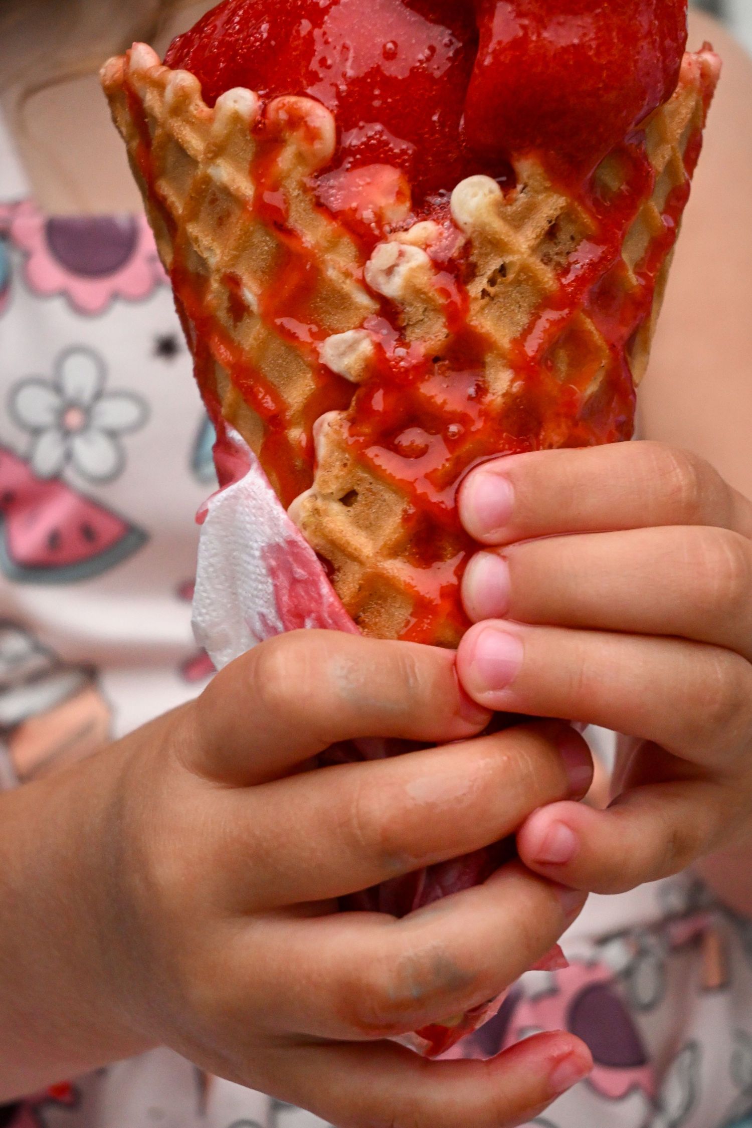 Child holds a large strawberry waffle cone ice cream with both hands.