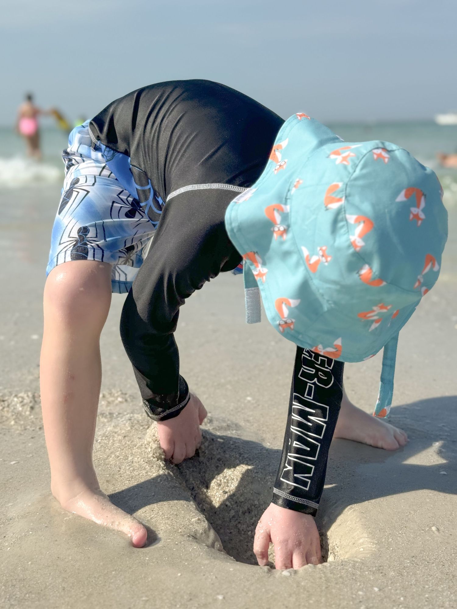 Young child in swimwear and teal hat digging in the sand at a sunny beach.