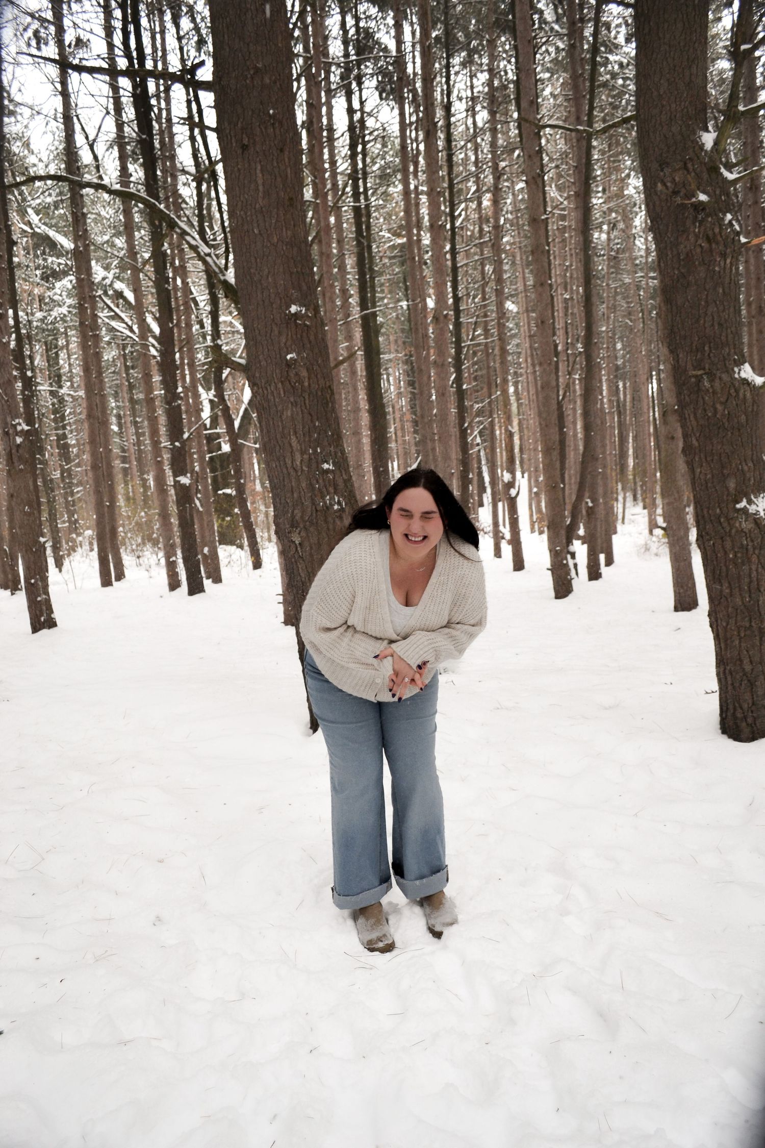 Happy woman laughing joyfully in a snowy pine forest wearing jeans and a cream sweater.
