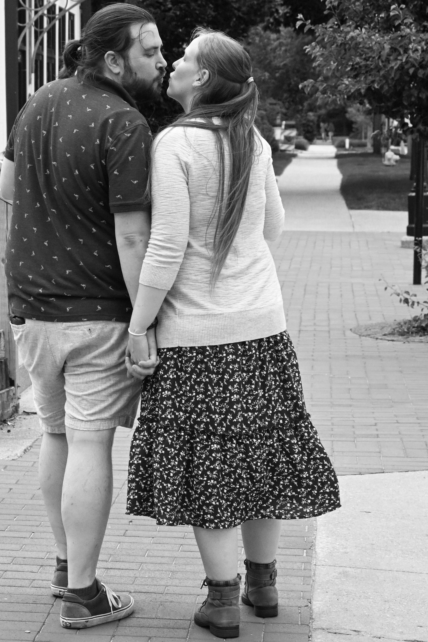 A black and white photo of a couple kissing while holding hands on a sidewalk.