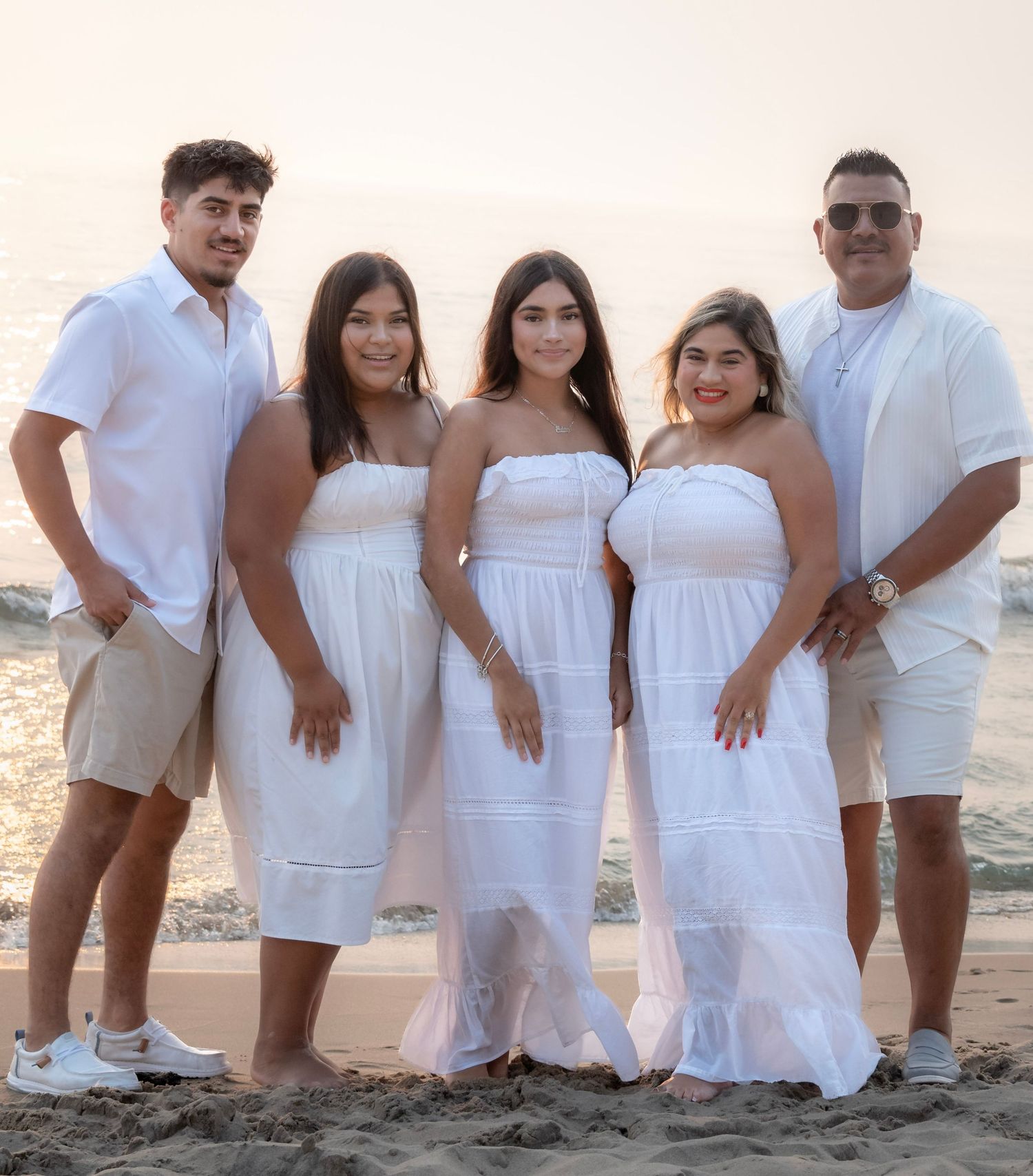 Family of five posing together on a beach at sunset, all wearing coordinated white outfits.