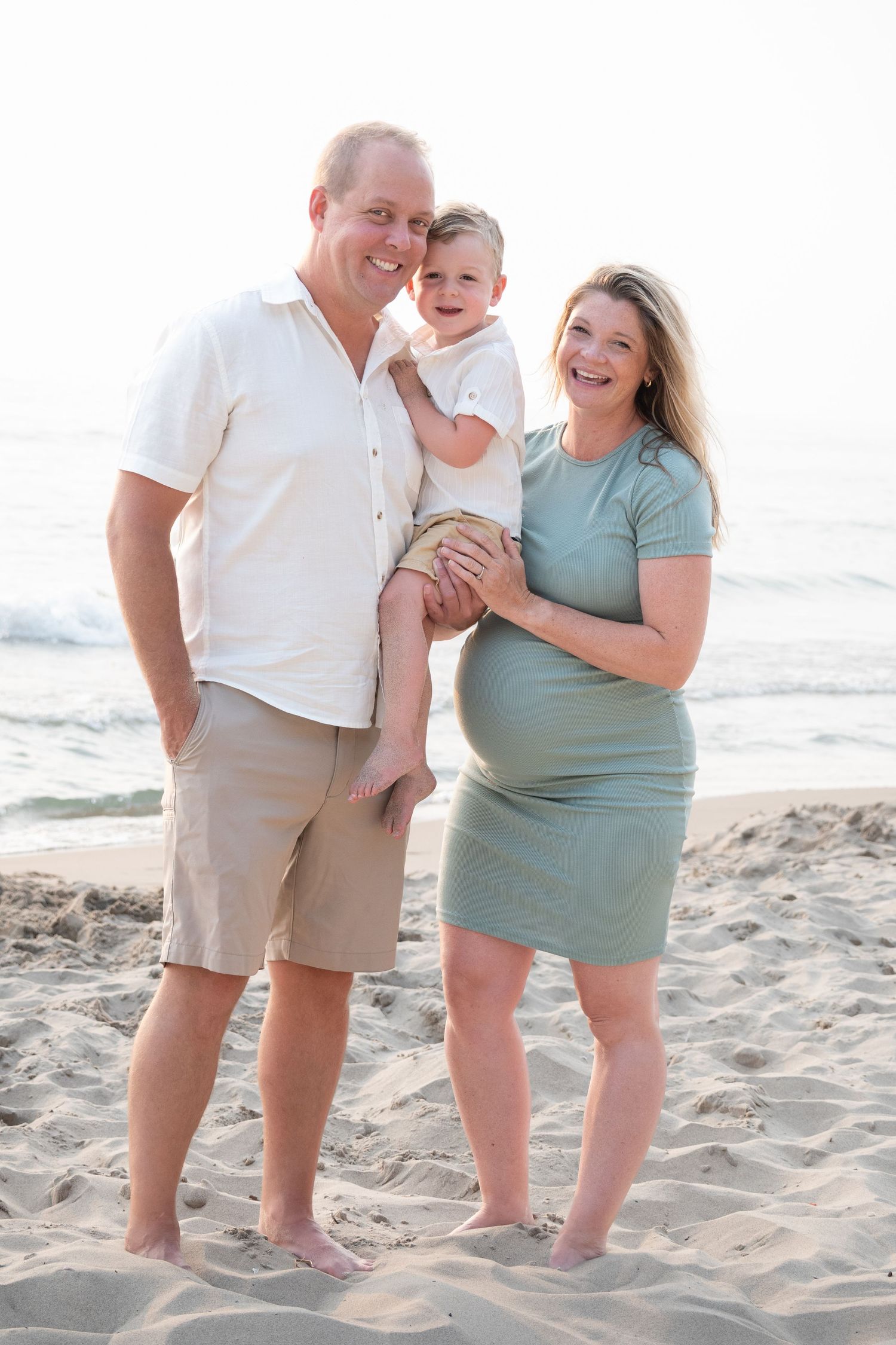 Smiling pregnant couple holding toddler on sandy beach, wearing casual summer outfits with ocean waves in background.