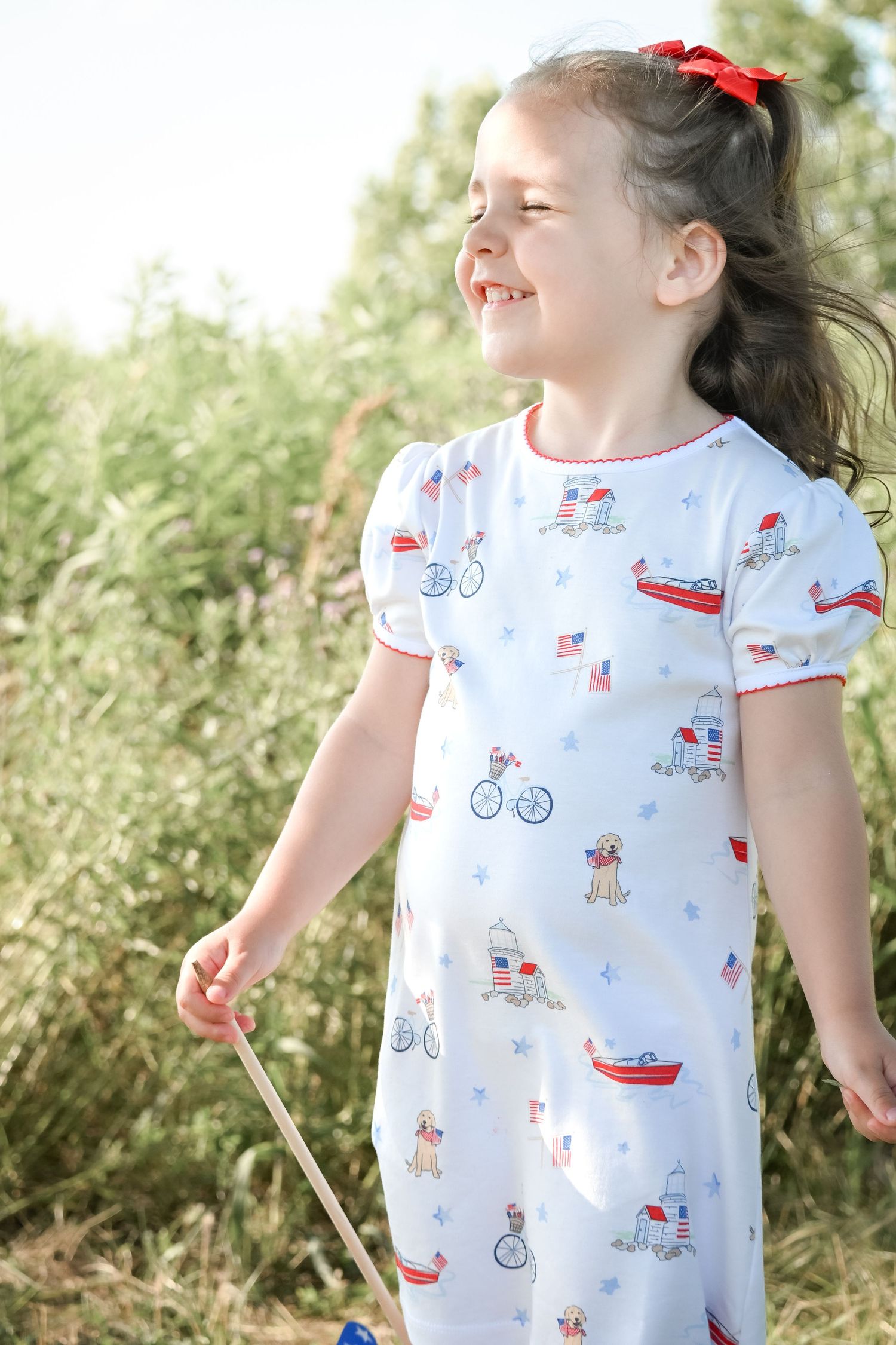 Young girl in patriotic print dress with red trim smiles outdoors, holding a stick near green foliage.