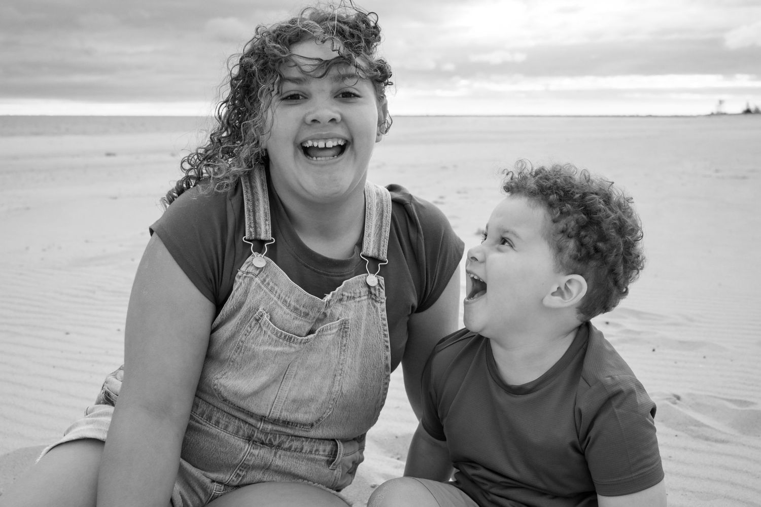 Black and white photo of two laughing siblings sitting together on a sandy beach.
