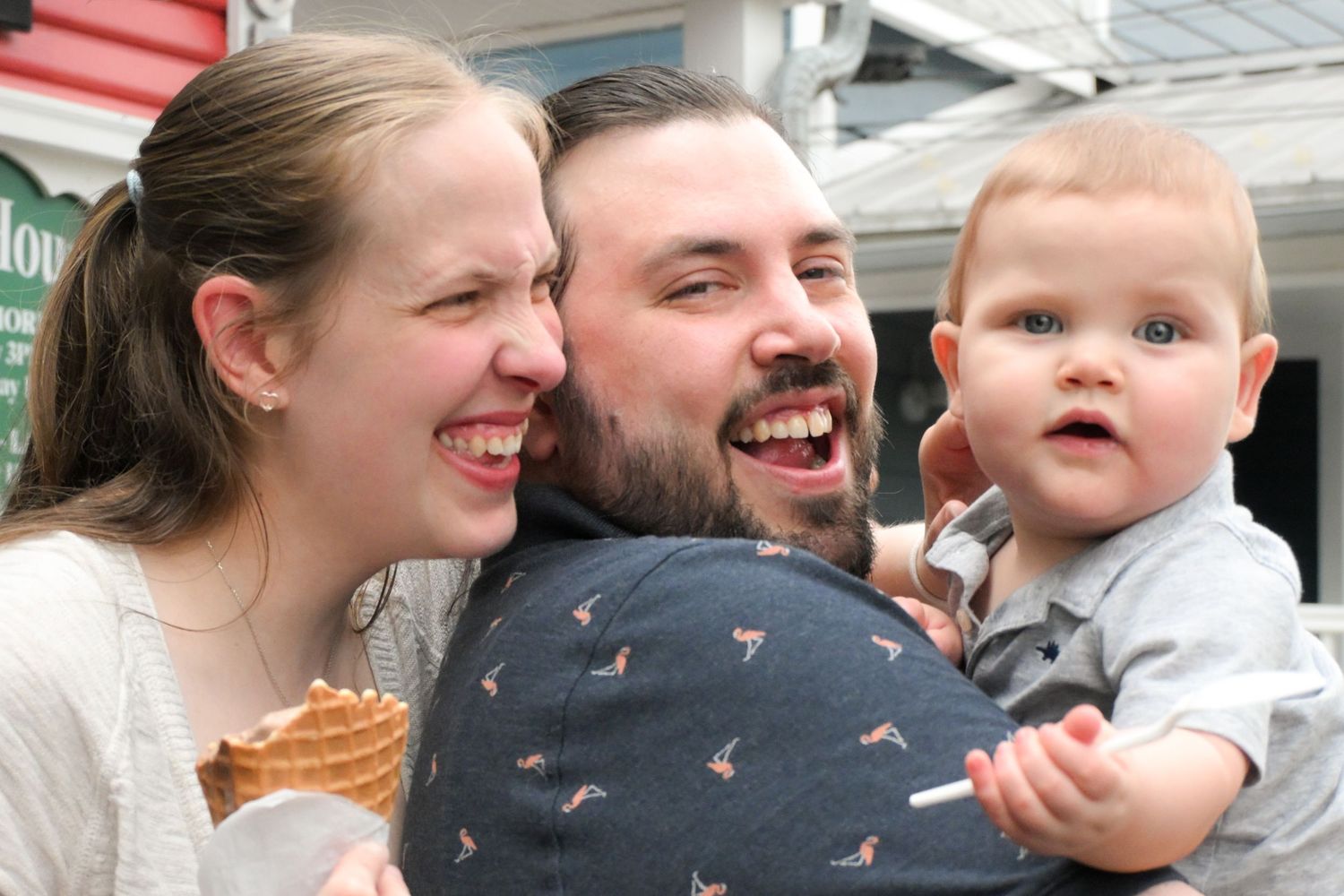 A happy couple laughs while holding an adorable wide-eyed baby outdoors near an ice cream shop.