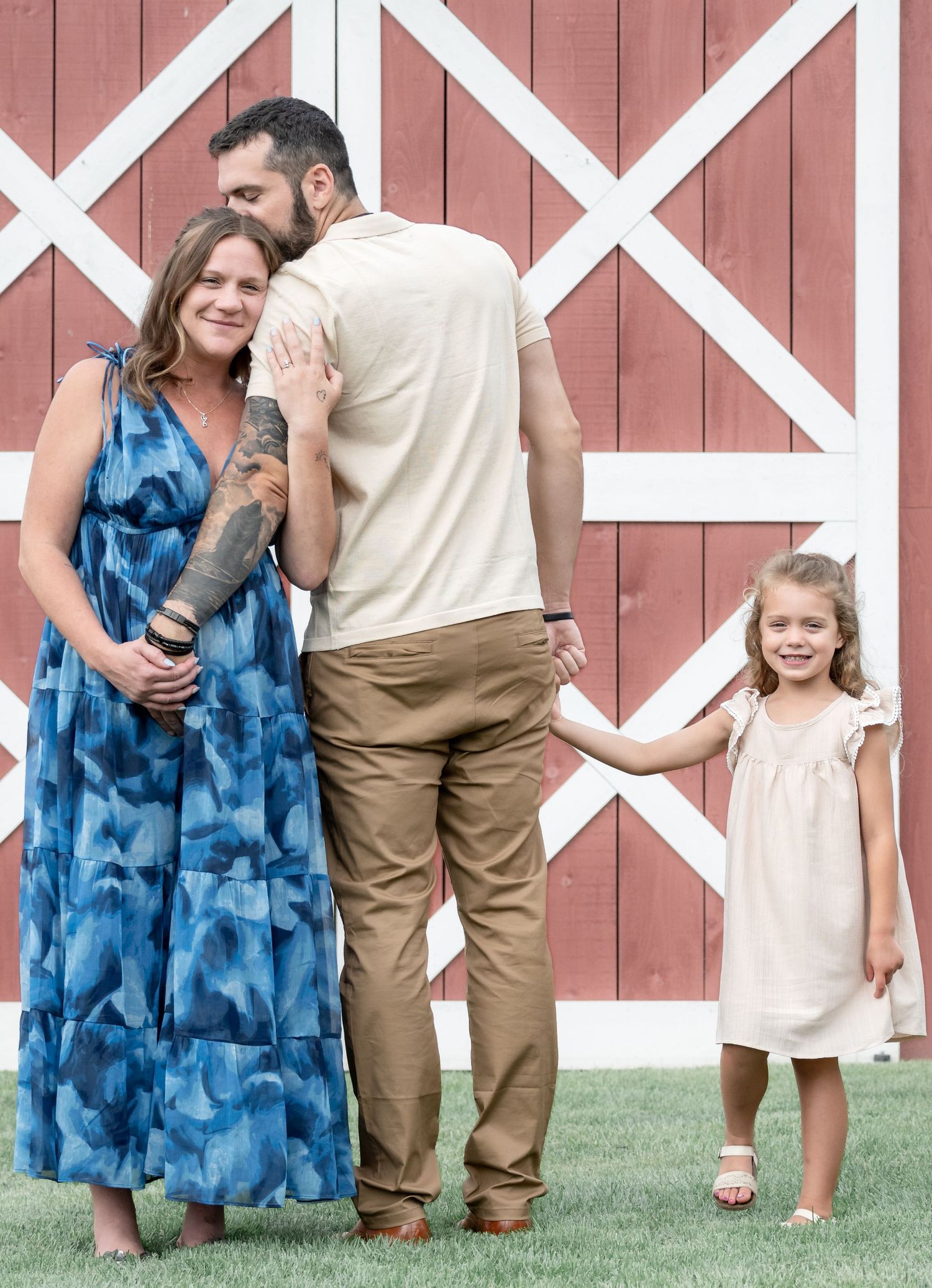 Family of three posing in front of red barn, woman in blue maxi dress, man in tan outfit, young girl in white dress.