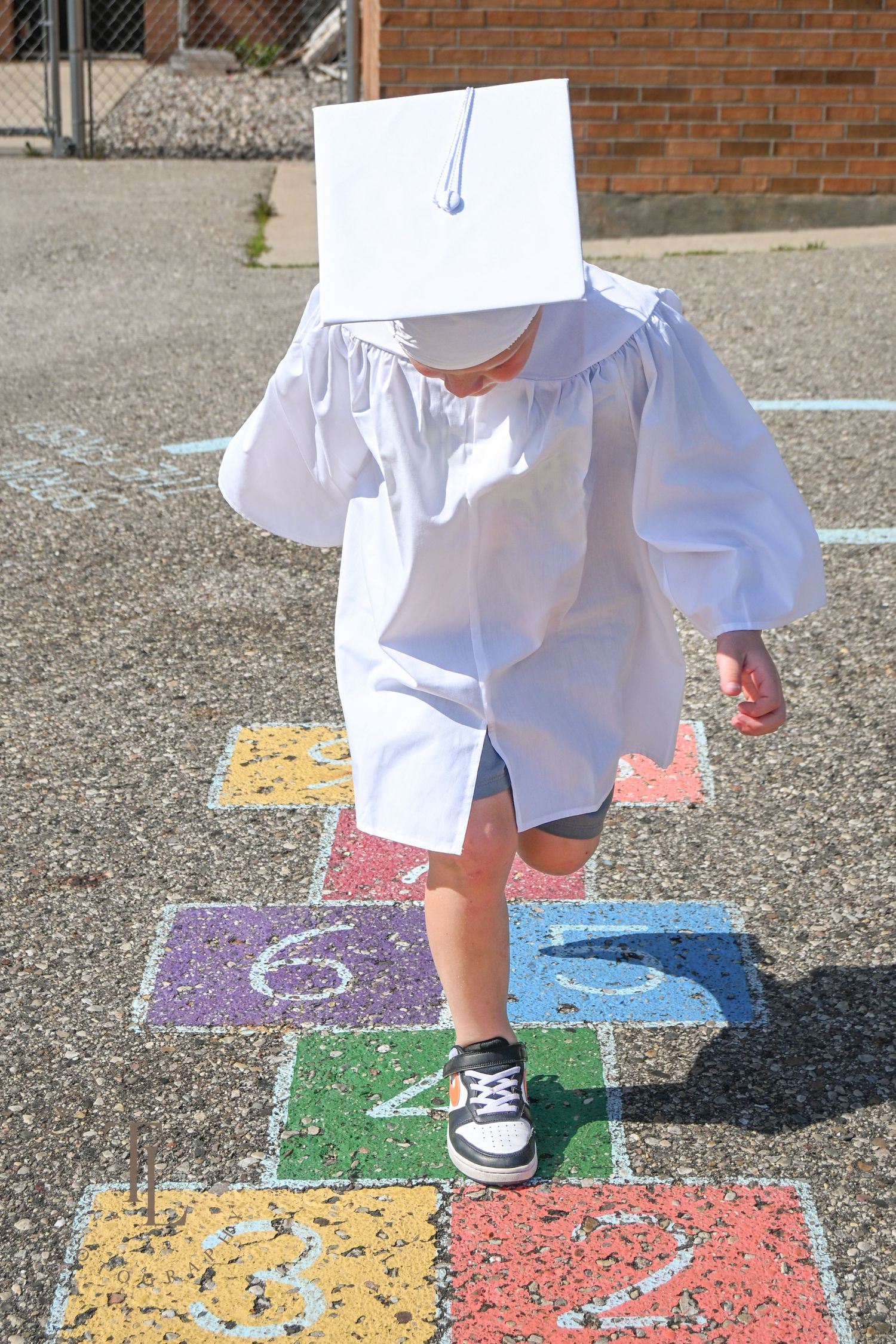 Young child in white graduation cap and gown playing hopscotch on a colorful school playground.