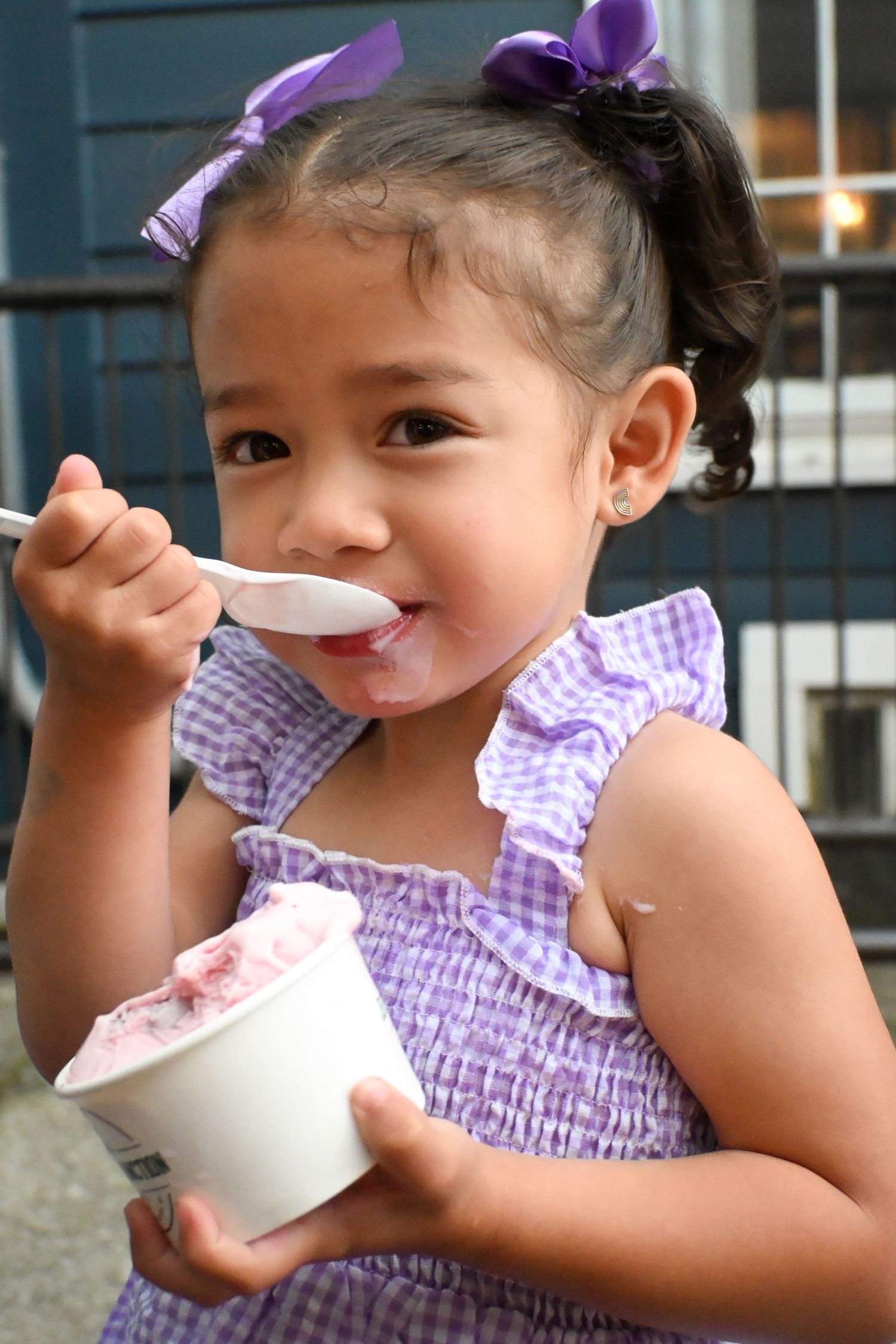 A young girl in a purple gingham dress eating pink ice cream from a cup with a spoon.