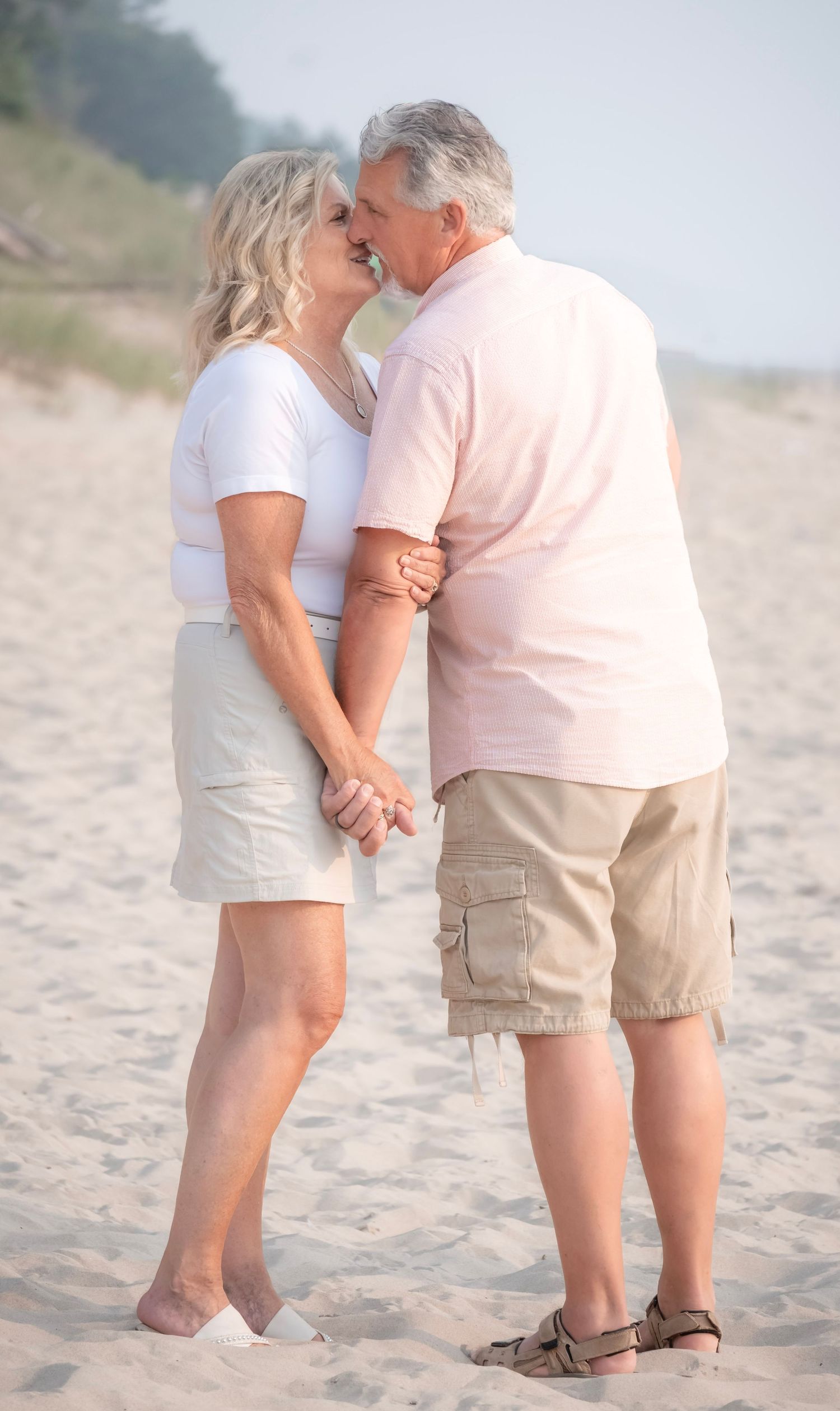 An older couple sharing a romantic kiss on a sandy beach, holding hands, dressed in casual summer attire.