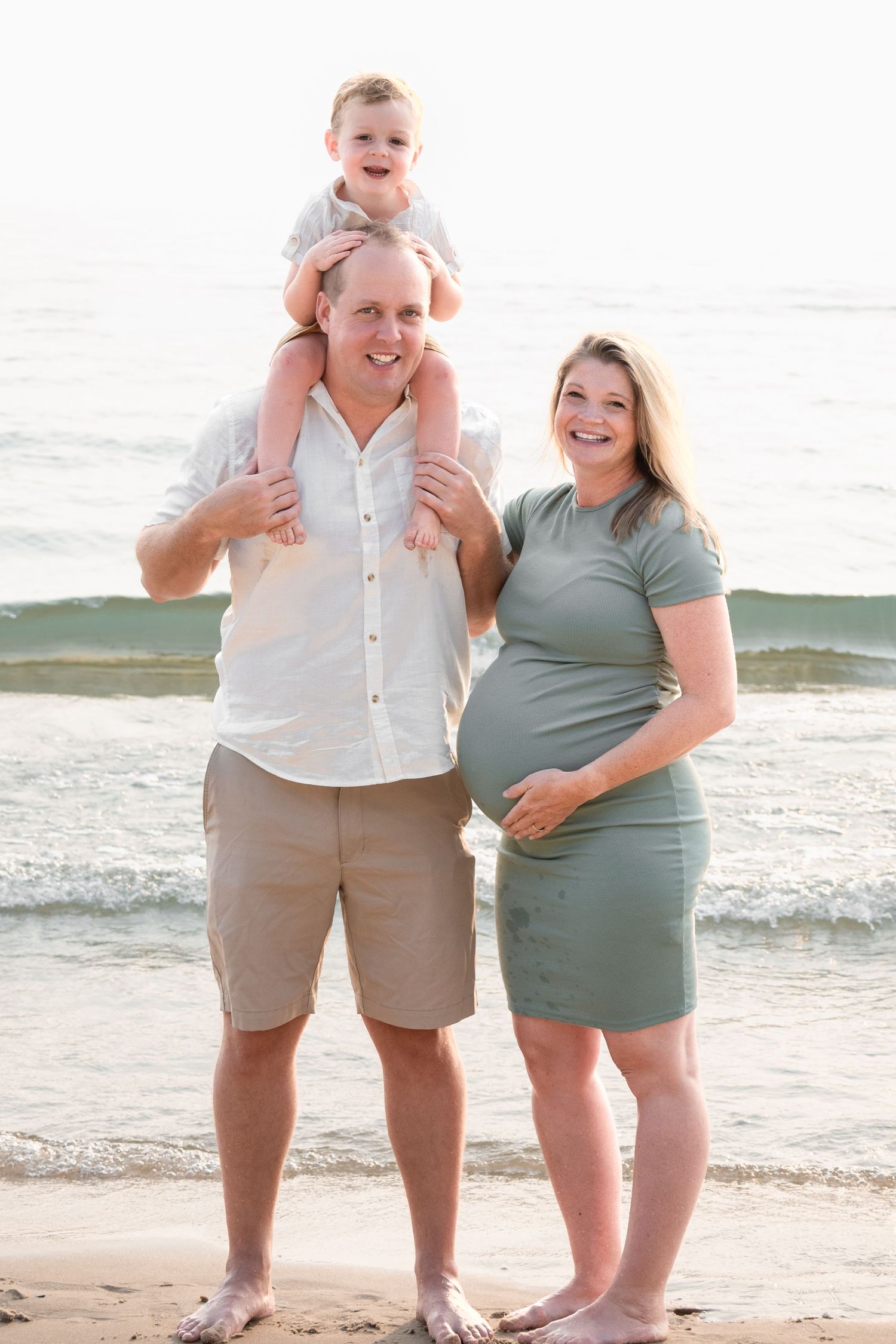 Pregnant woman in sage dress poses with husband and toddler on shoulders at beach.