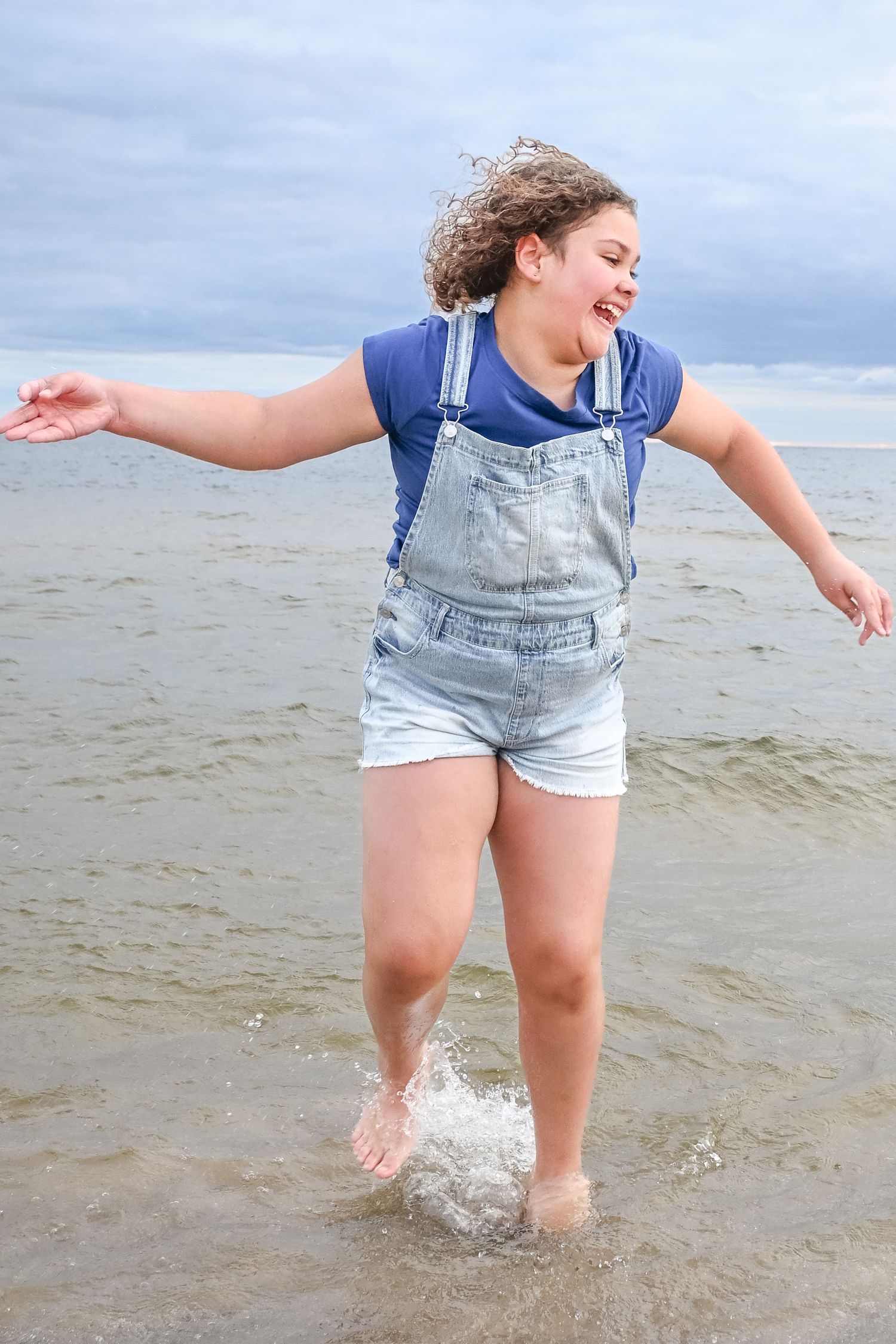 A laughing girl in denim overalls splashes through shallow ocean waves at the beach.
