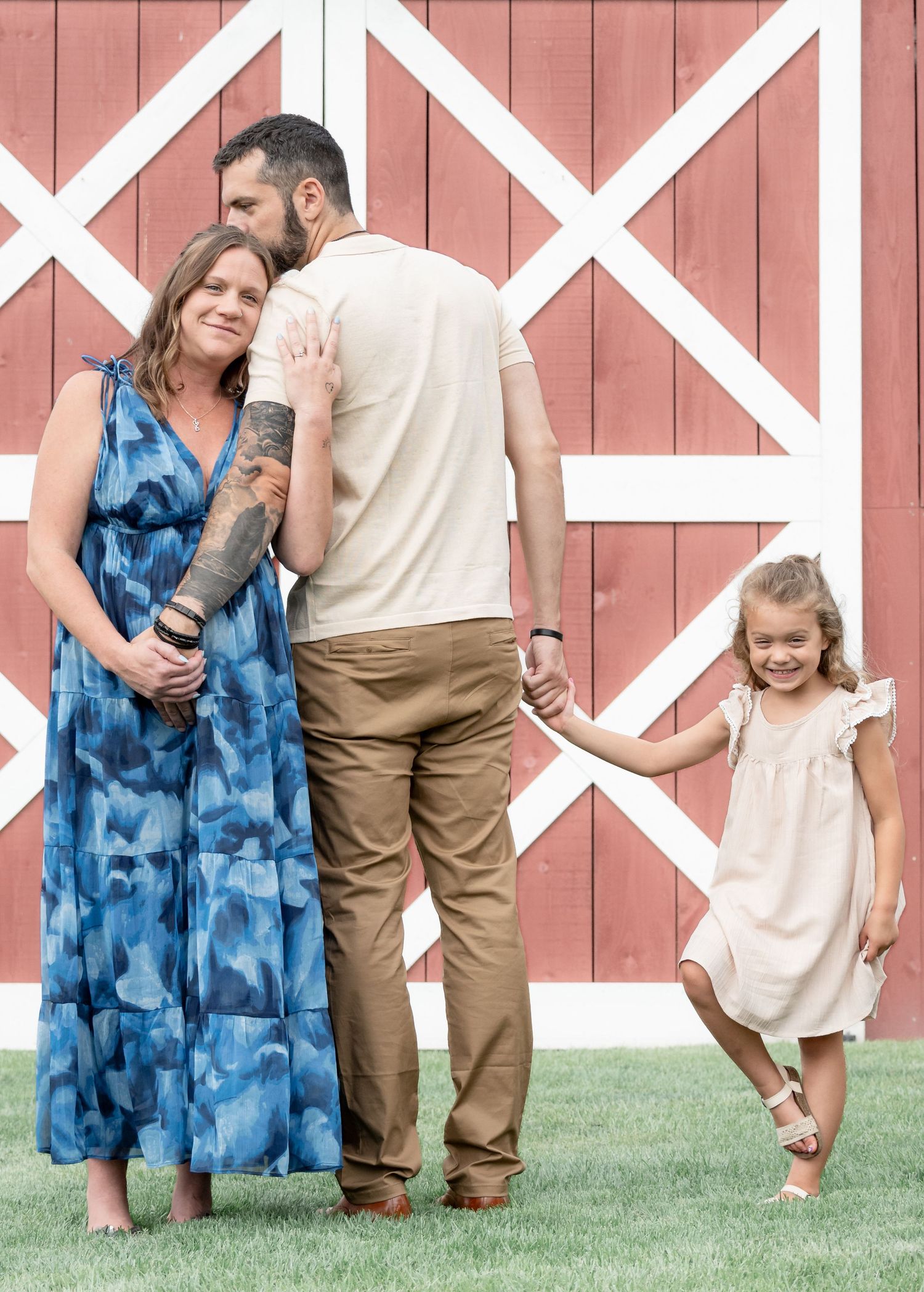 Family of three posing in front of a red barn, parents embracing while young daughter holds father's hand.
