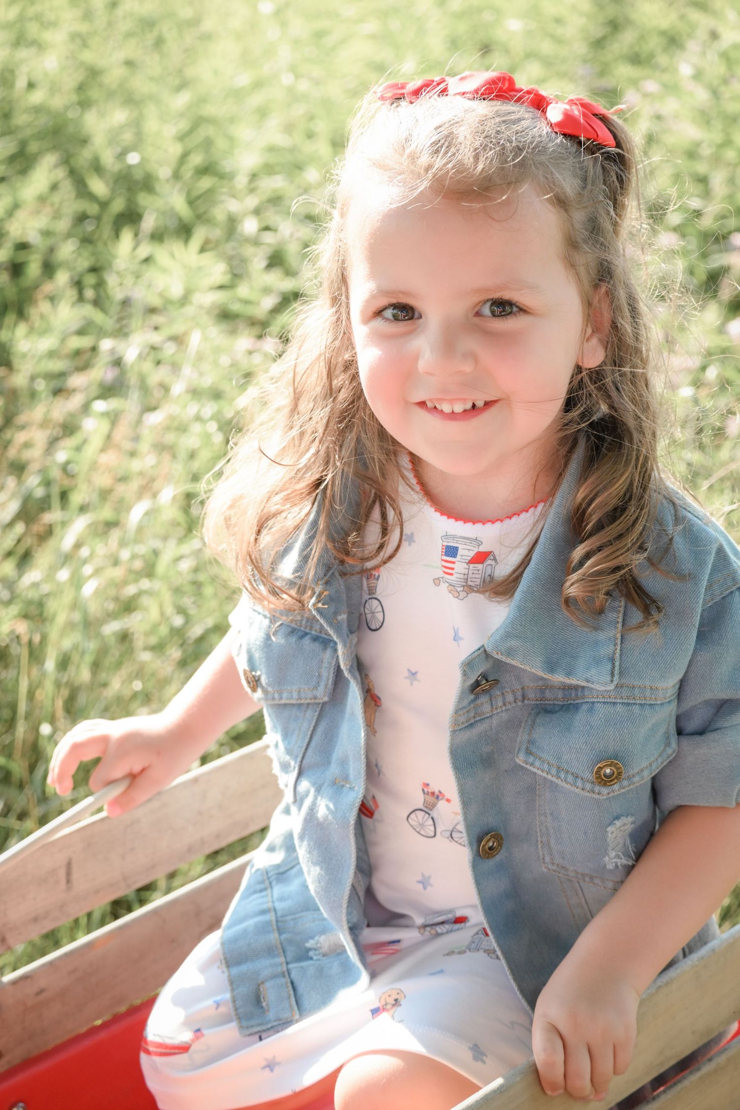 Smiling young girl in denim jacket and floral dress sitting in a red wagon outdoors on a sunny day.