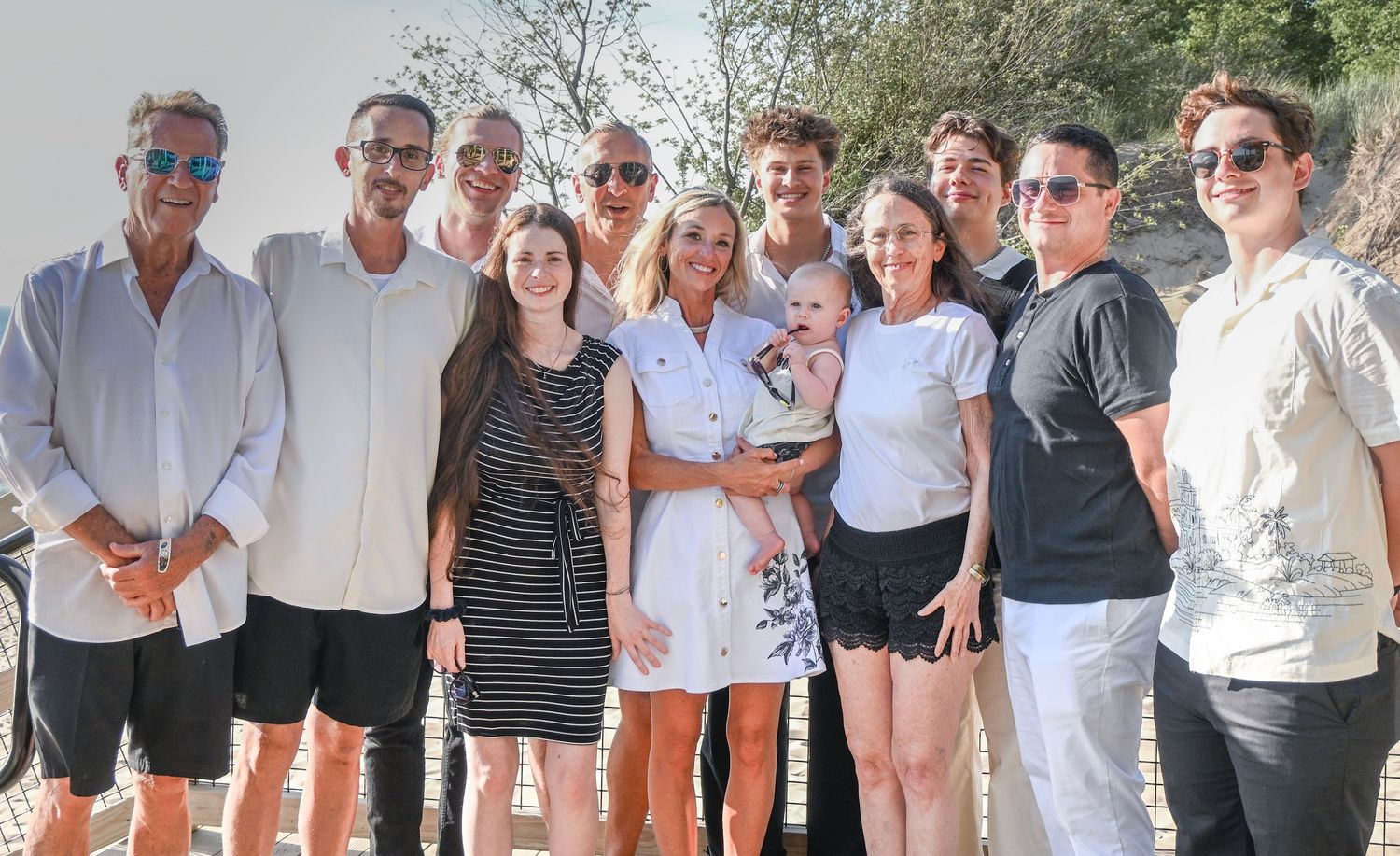 Large family group posing together outdoors on a sunny day, smiling for a photo with trees in the background.