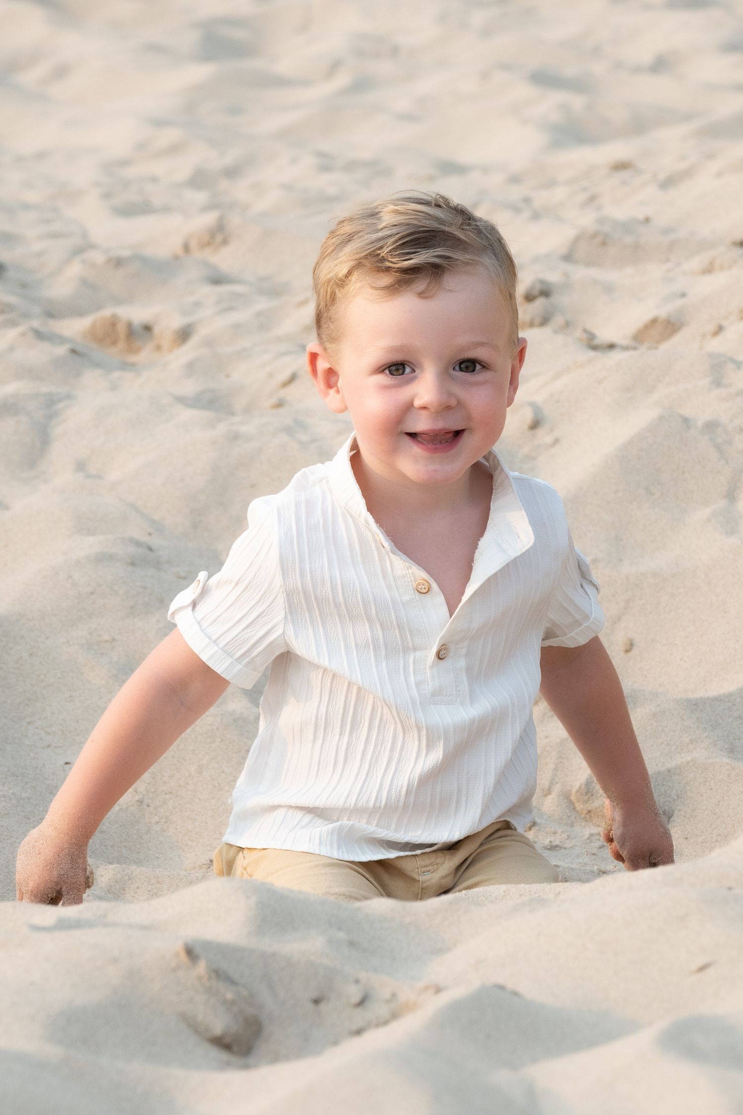 A happy toddler boy in a white shirt and tan shorts smiles while playing in the sand at the beach.