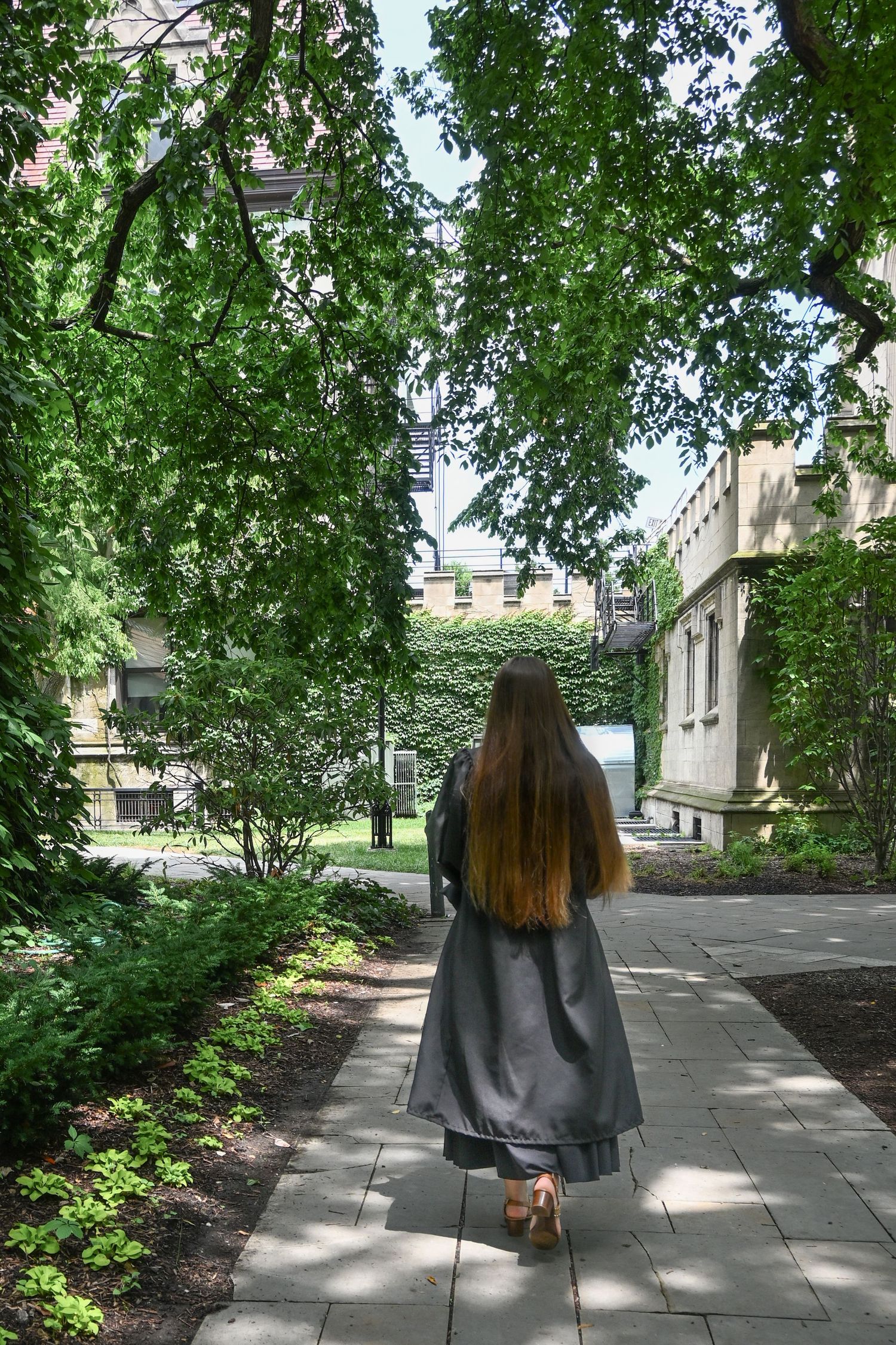 A woman in a gray graduation gown walks along a tree-lined campus pathway surrounded by lush green foliage.
