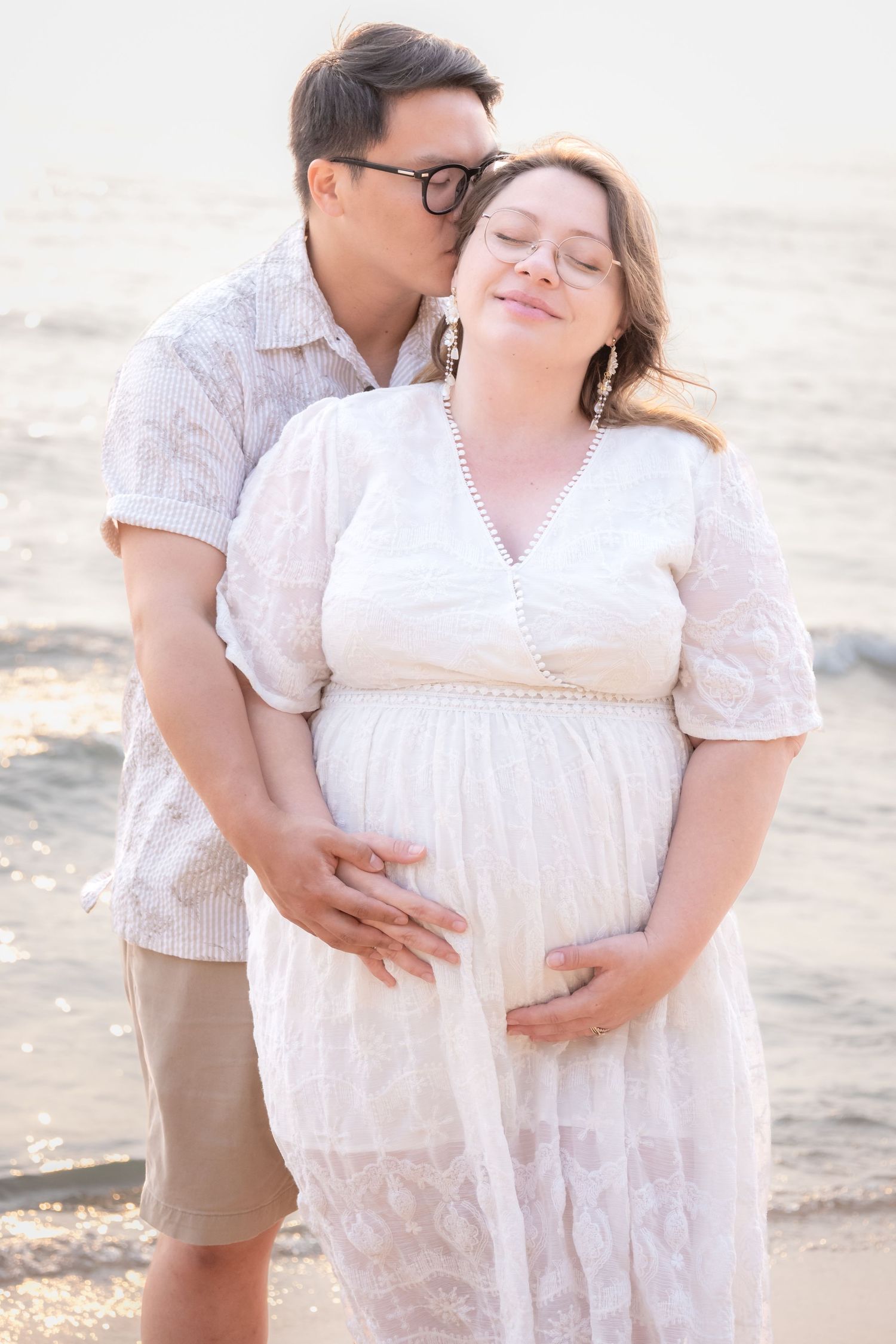 A man kisses a pregnant woman on the cheek while holding her belly on a sunny beach.