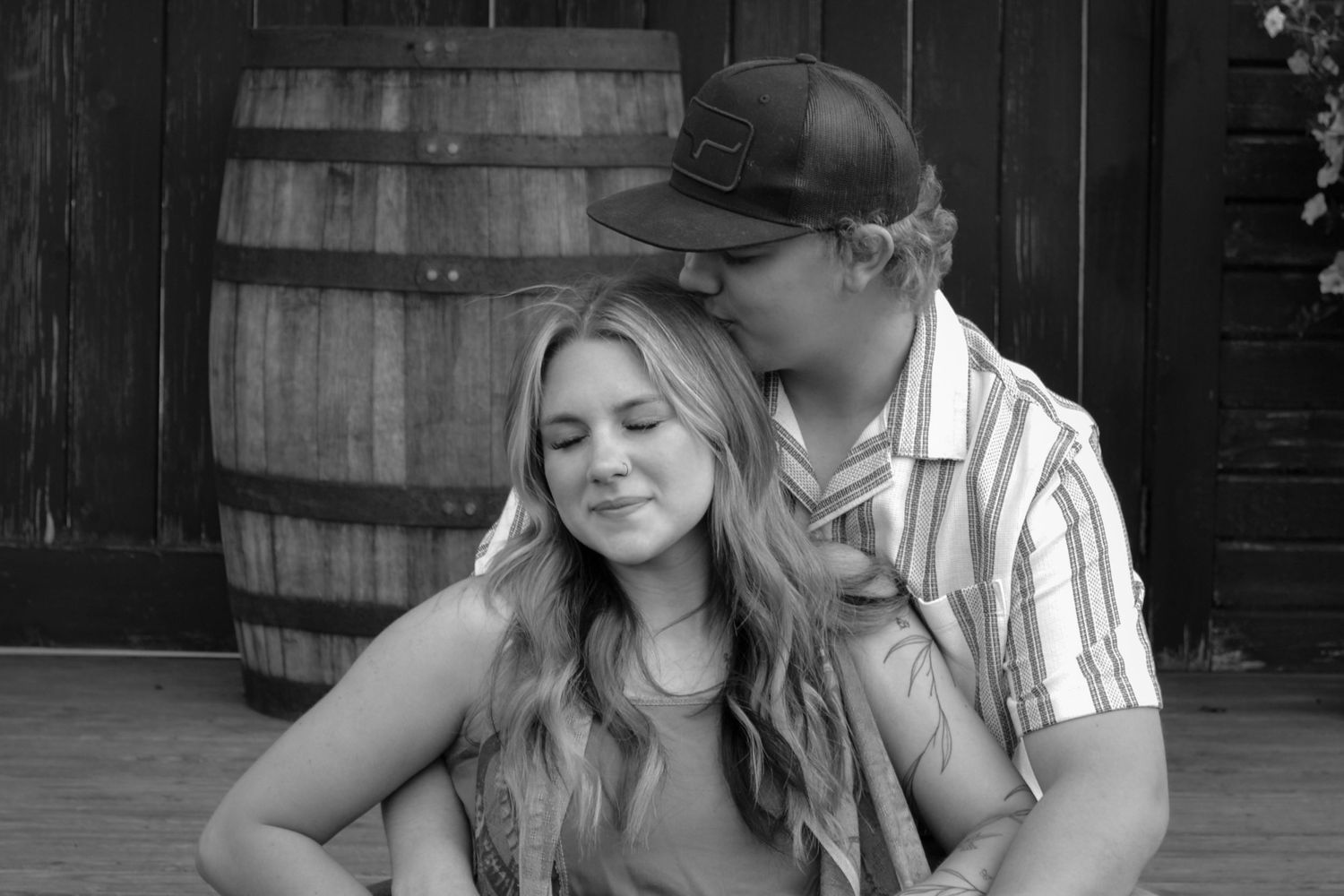 A young couple sitting together near a wooden barrel in a black and white romantic portrait photo.