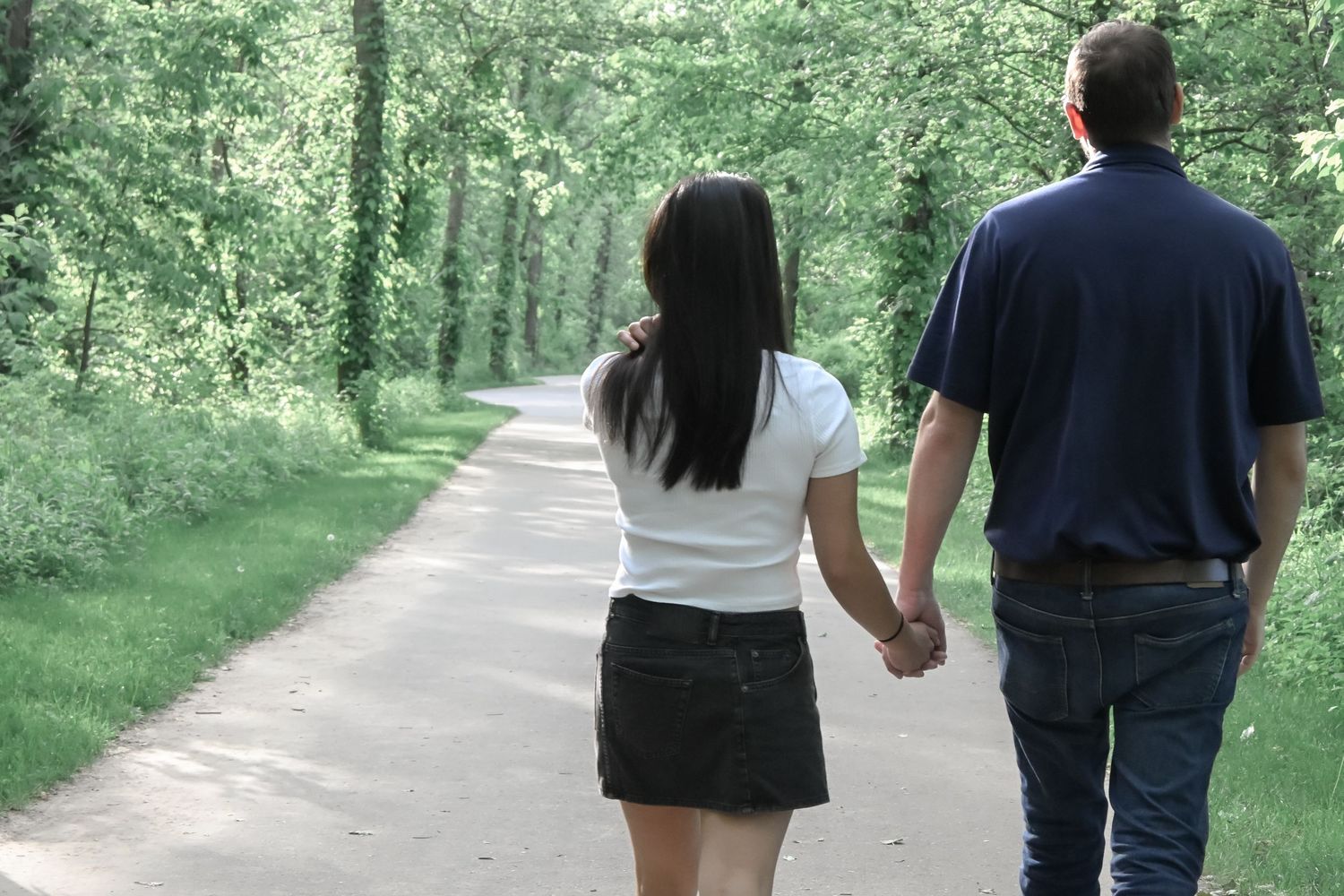 Couple holding hands walking down a tree-lined path in a lush green park on a peaceful day.