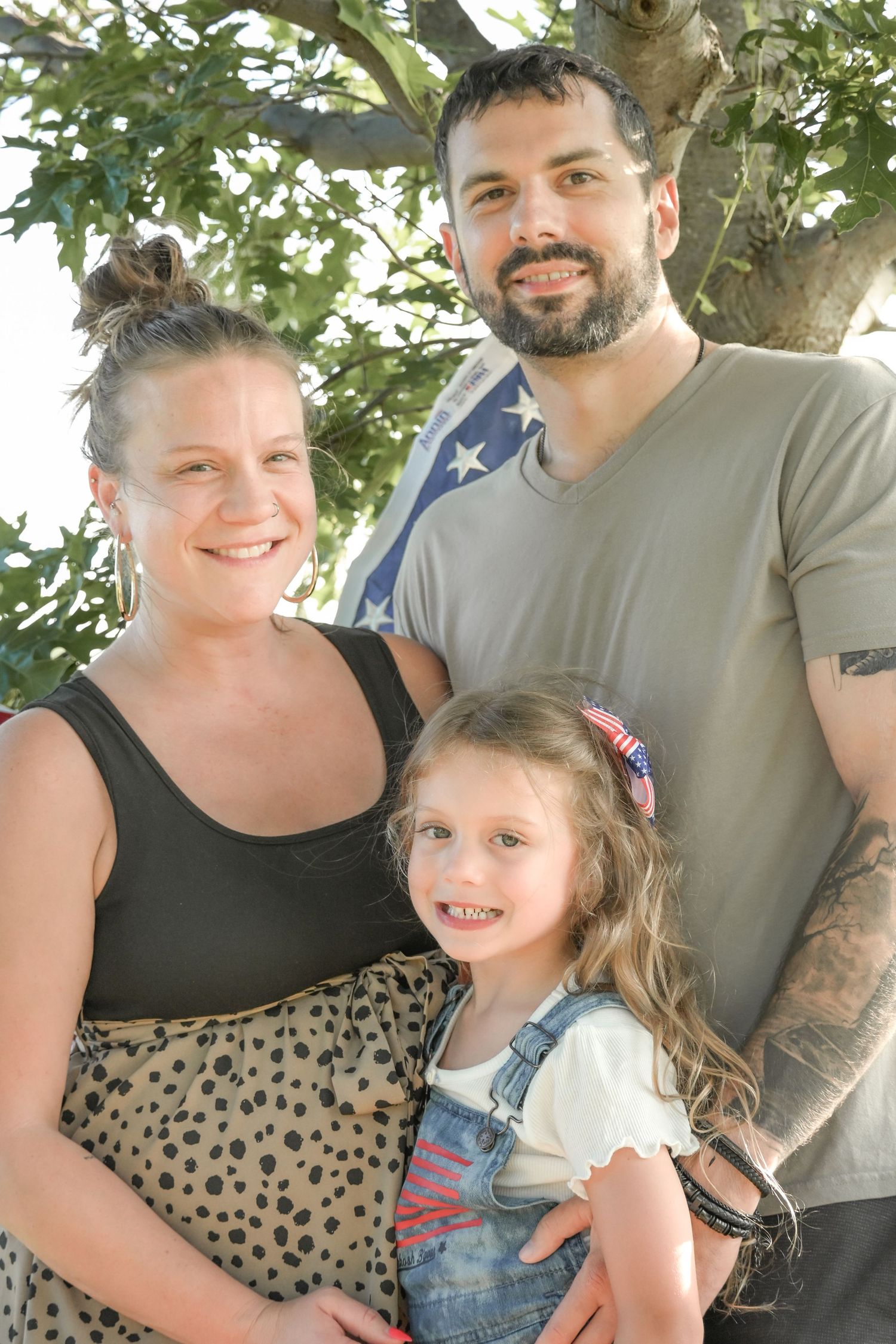 Family of three smiling outdoors under a tree with an American flag, mom, dad, and young girl in denim overalls.