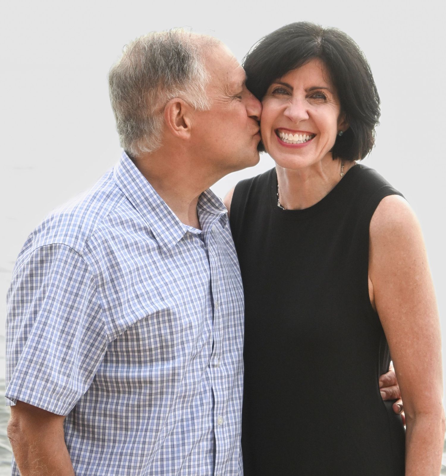 An older man kisses a smiling woman on the cheek as they pose together outdoors.