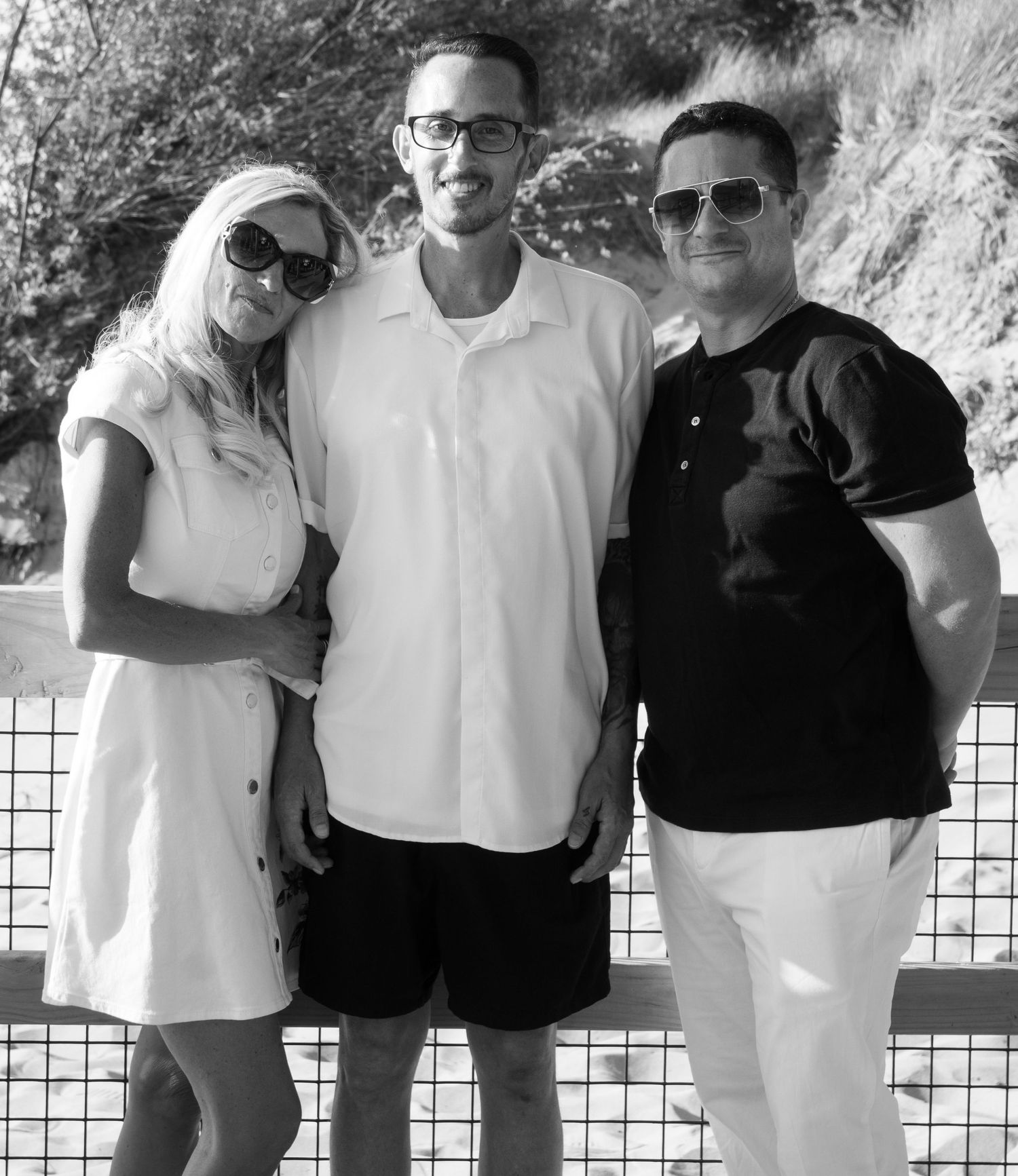 Black and white photo of three smiling adults posing together outdoors, wearing sunglasses on a sunny day.