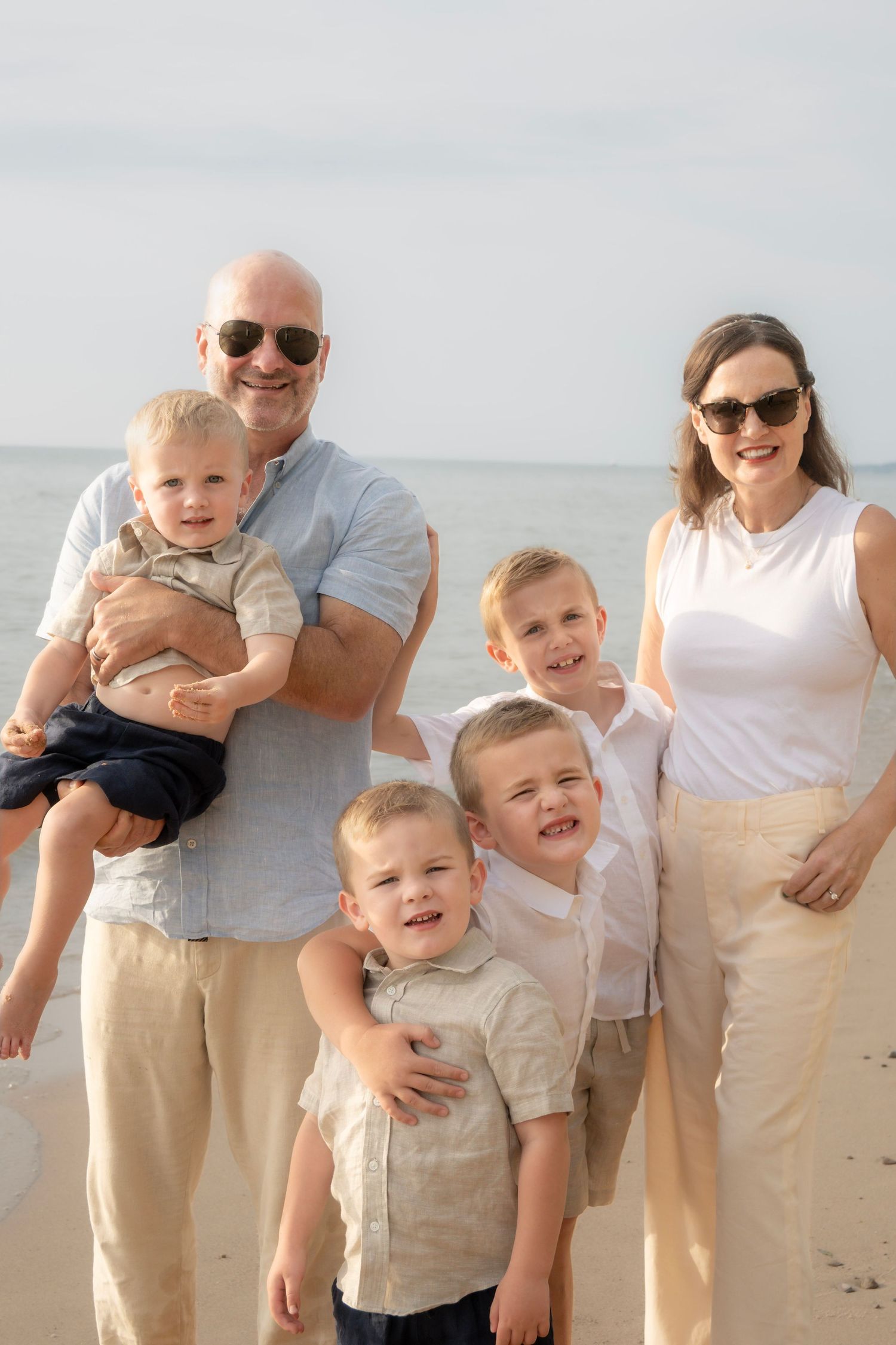 Family of six posing on a beach, parents with four young boys, dressed in neutral tones on a sunny day.