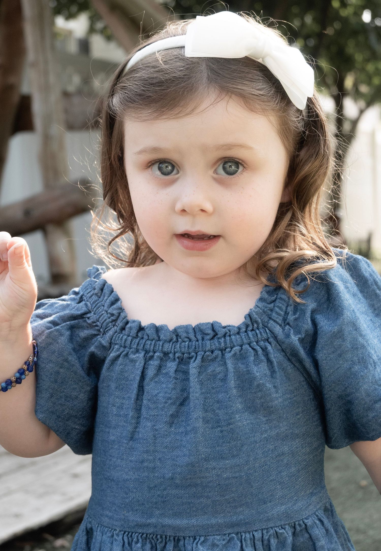 Adorable toddler girl with blue eyes wearing a denim off-shoulder dress and white bow headband.