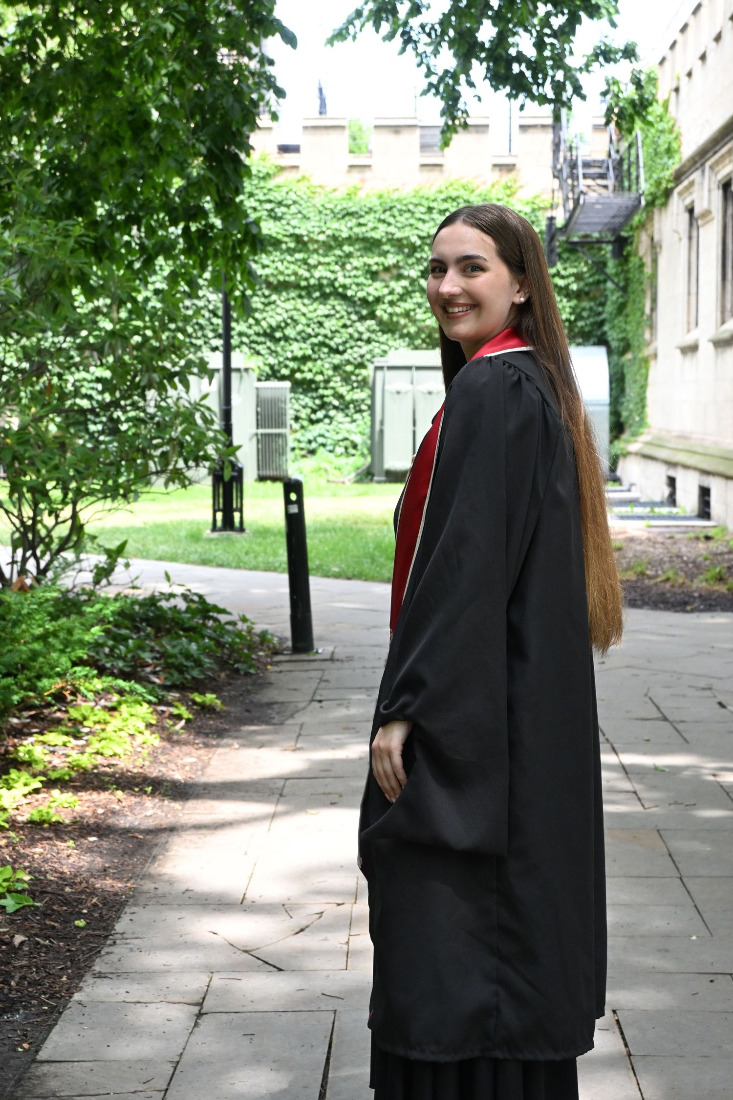 Young woman in black graduation gown with red trim smiles over shoulder on a tree-lined campus pathway.