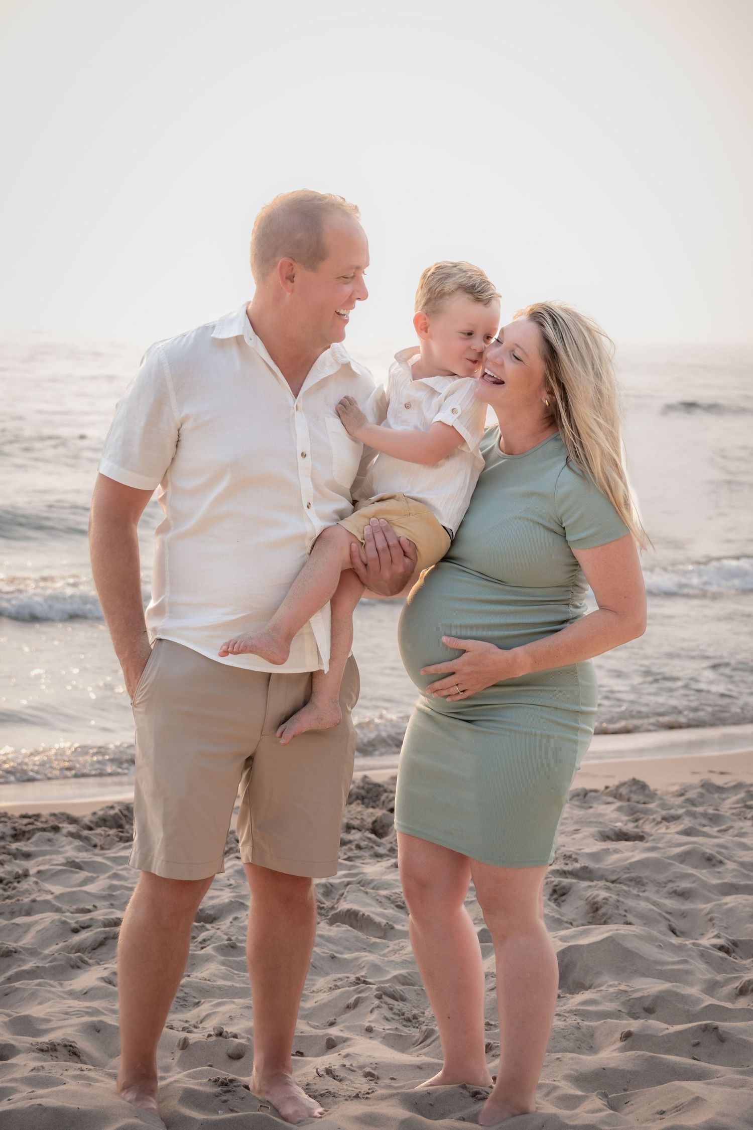 Pregnant woman in sage green dress with husband and toddler son smiling together on a sandy beach at sunset.