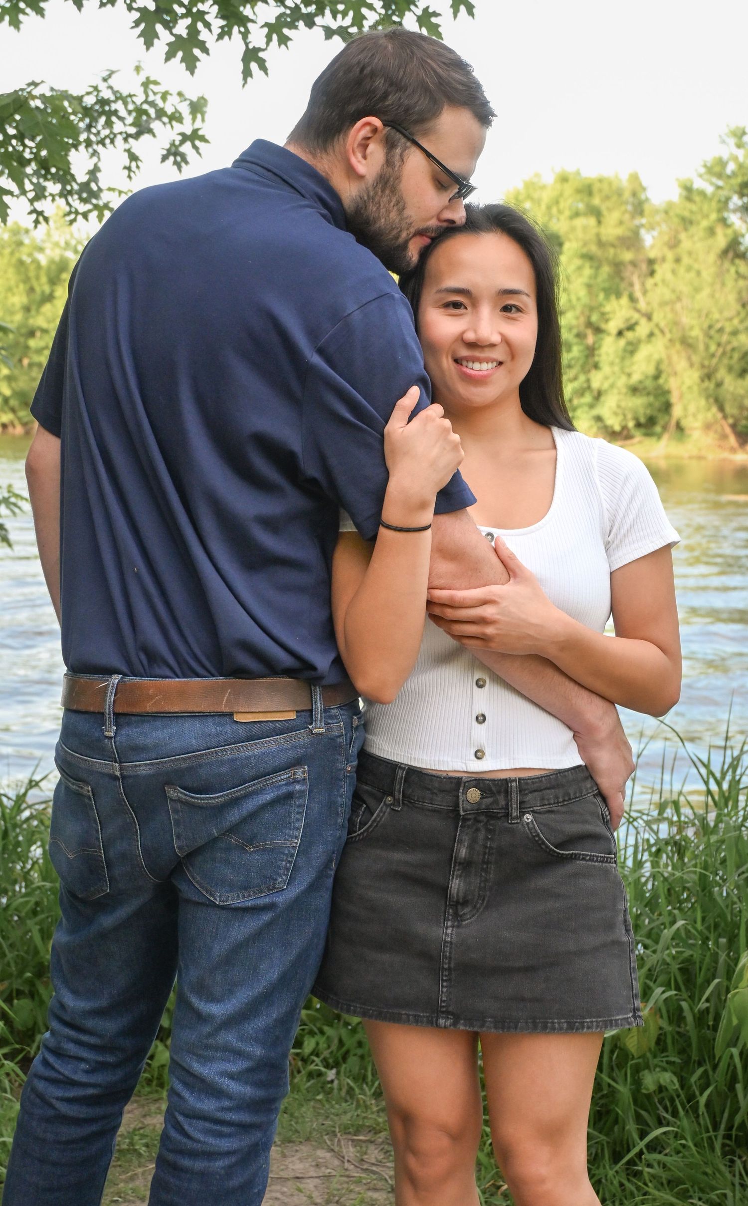 A couple shares a tender moment outdoors near a river, man kissing woman's cheek as she smiles warmly.