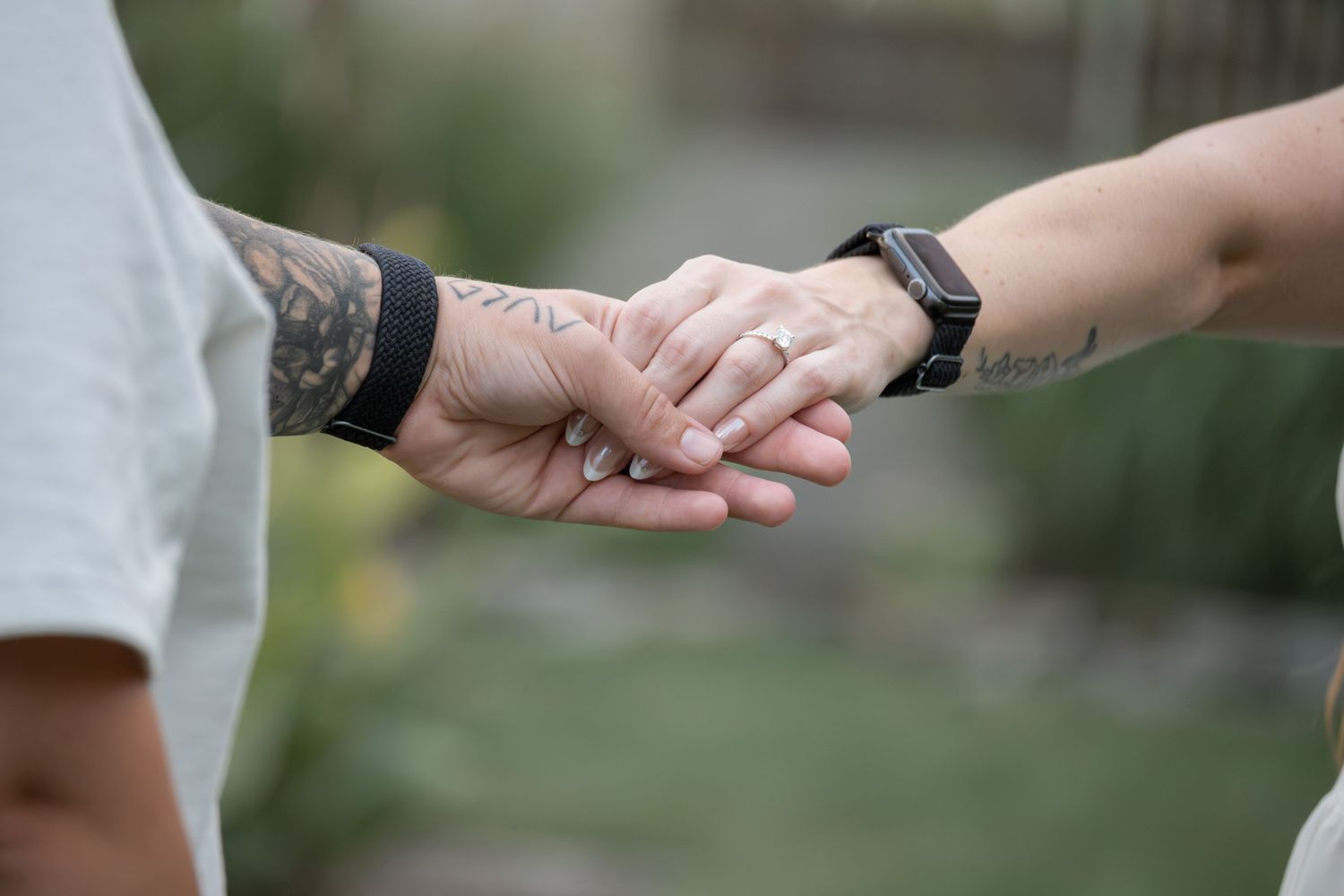 Two tattooed hands holding gently, one wearing a ring and smartwatch, captured outdoors with soft bokeh background.