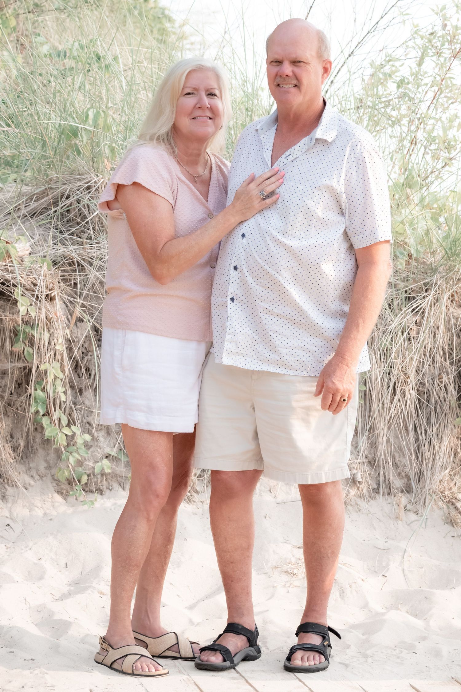 Happy senior couple posing together on sandy beach dunes, woman in pink top touching mans chest.