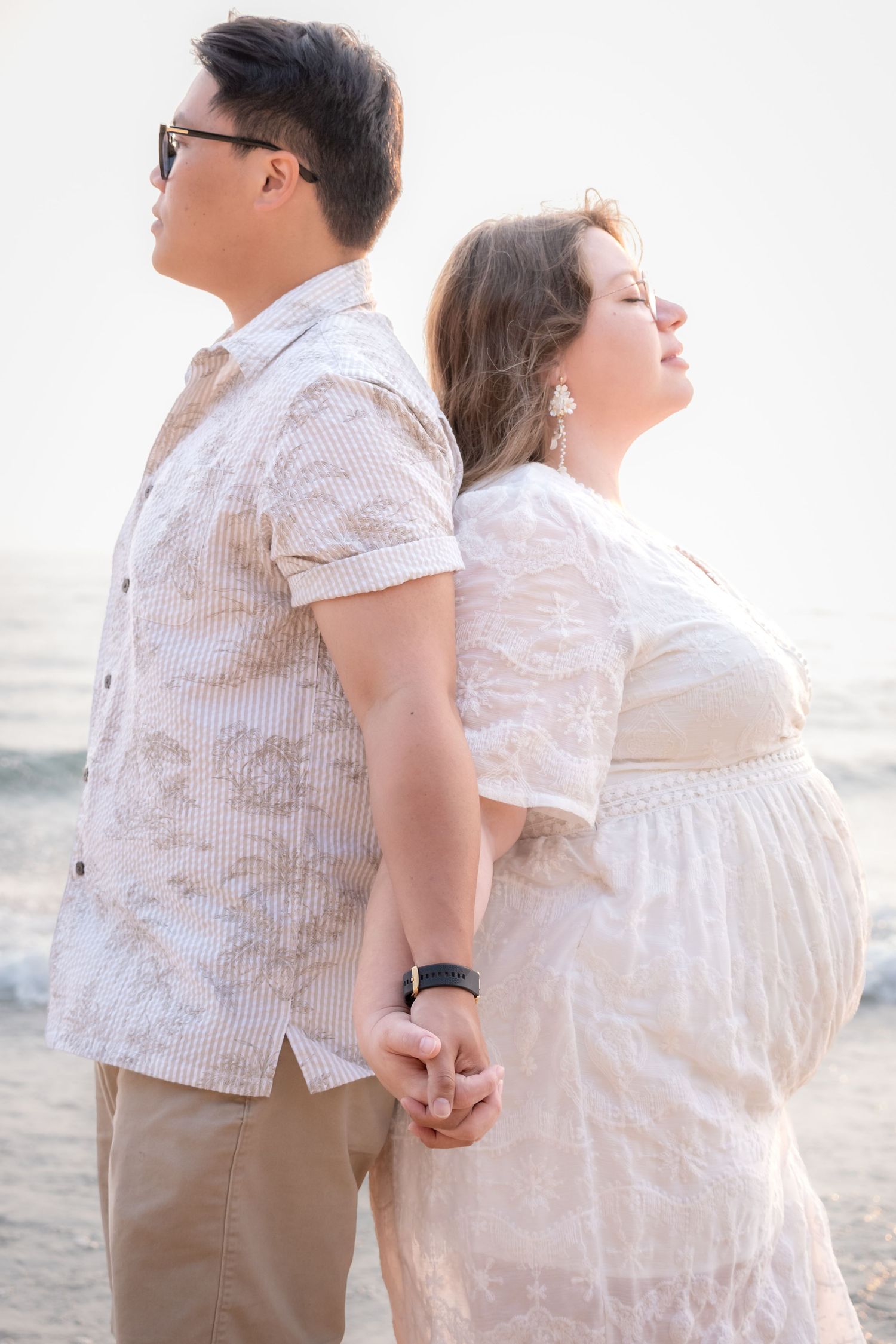 Pregnant couple holding hands back-to-back on a beach at sunset, wearing coordinating white and beige outfits.