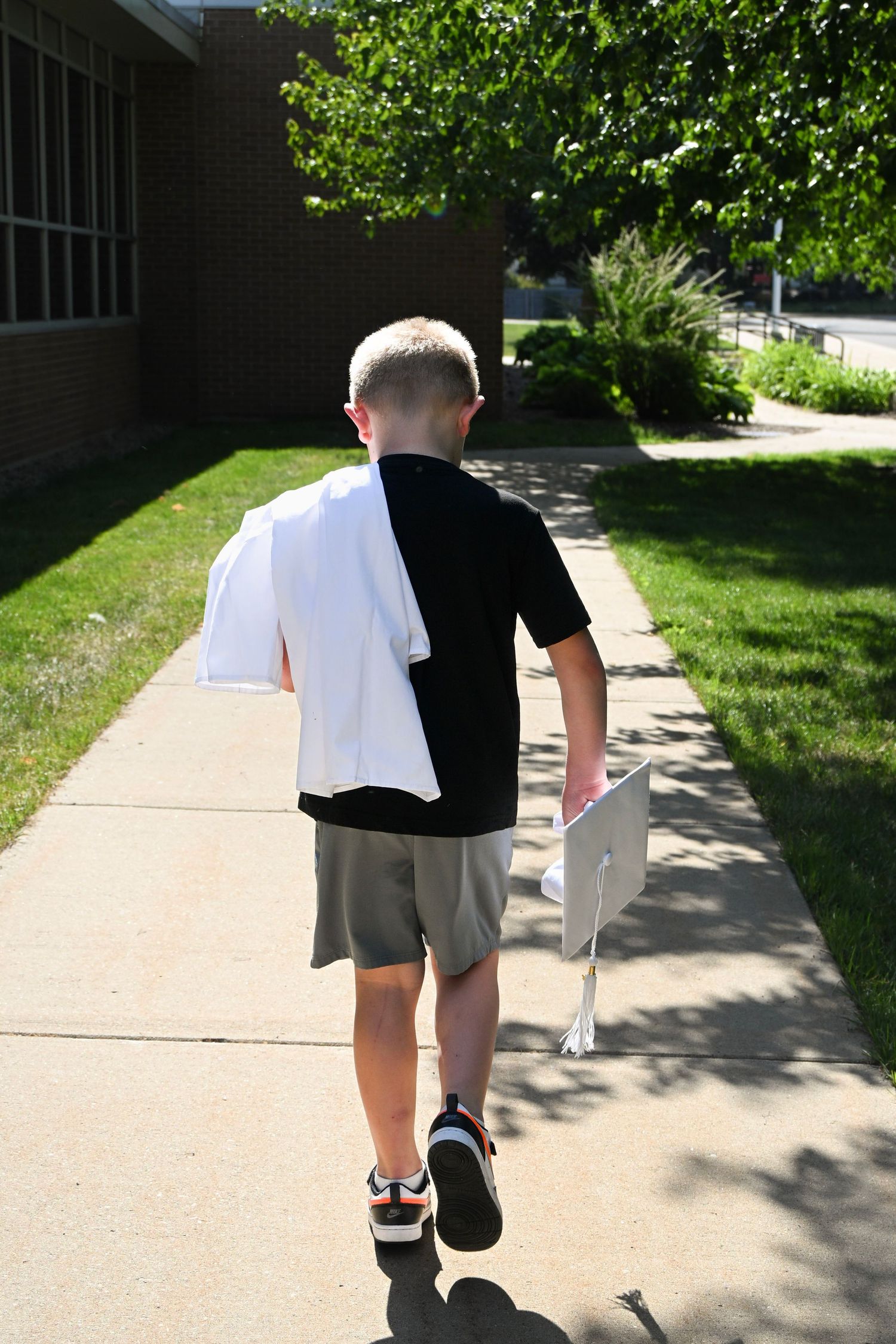 Boy walking on sidewalk carrying laptop and white shirt toward brick school building on sunny day.