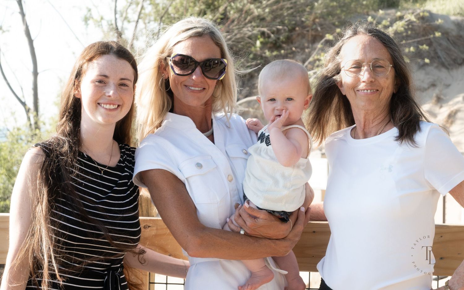Four generations of women pose outdoors, with one holding a baby, all dressed in white and stripes on a sunny day.