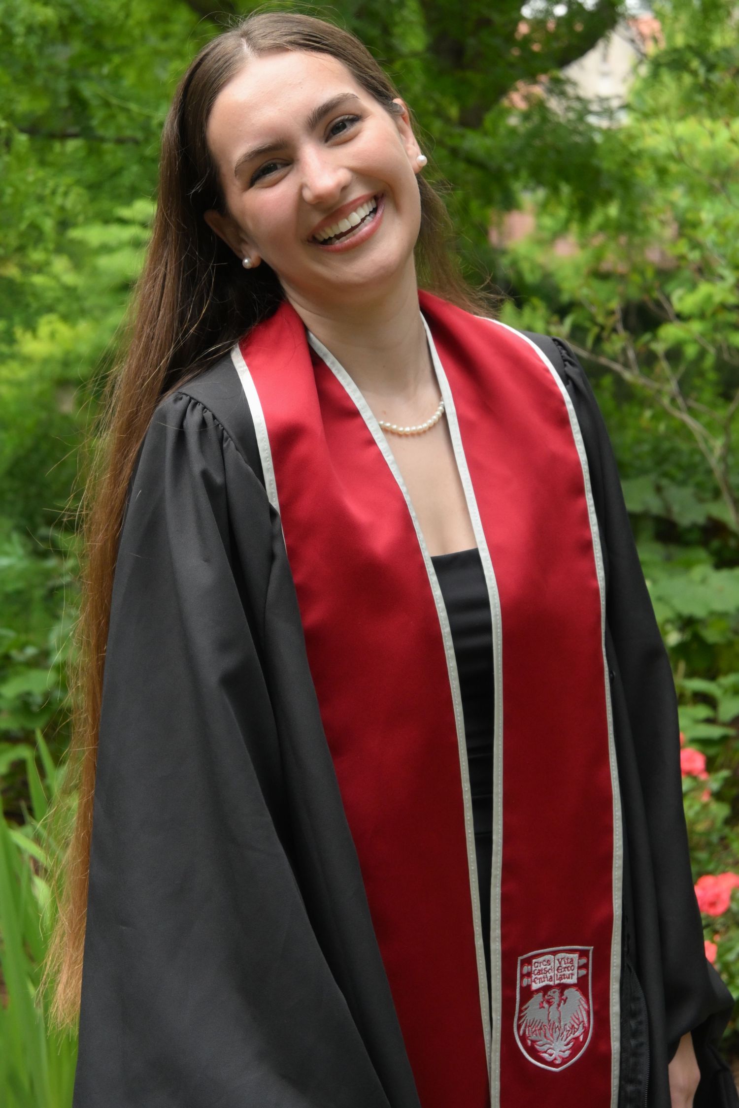 Happy young woman in black graduation gown and red sash smiling in lush green garden setting.