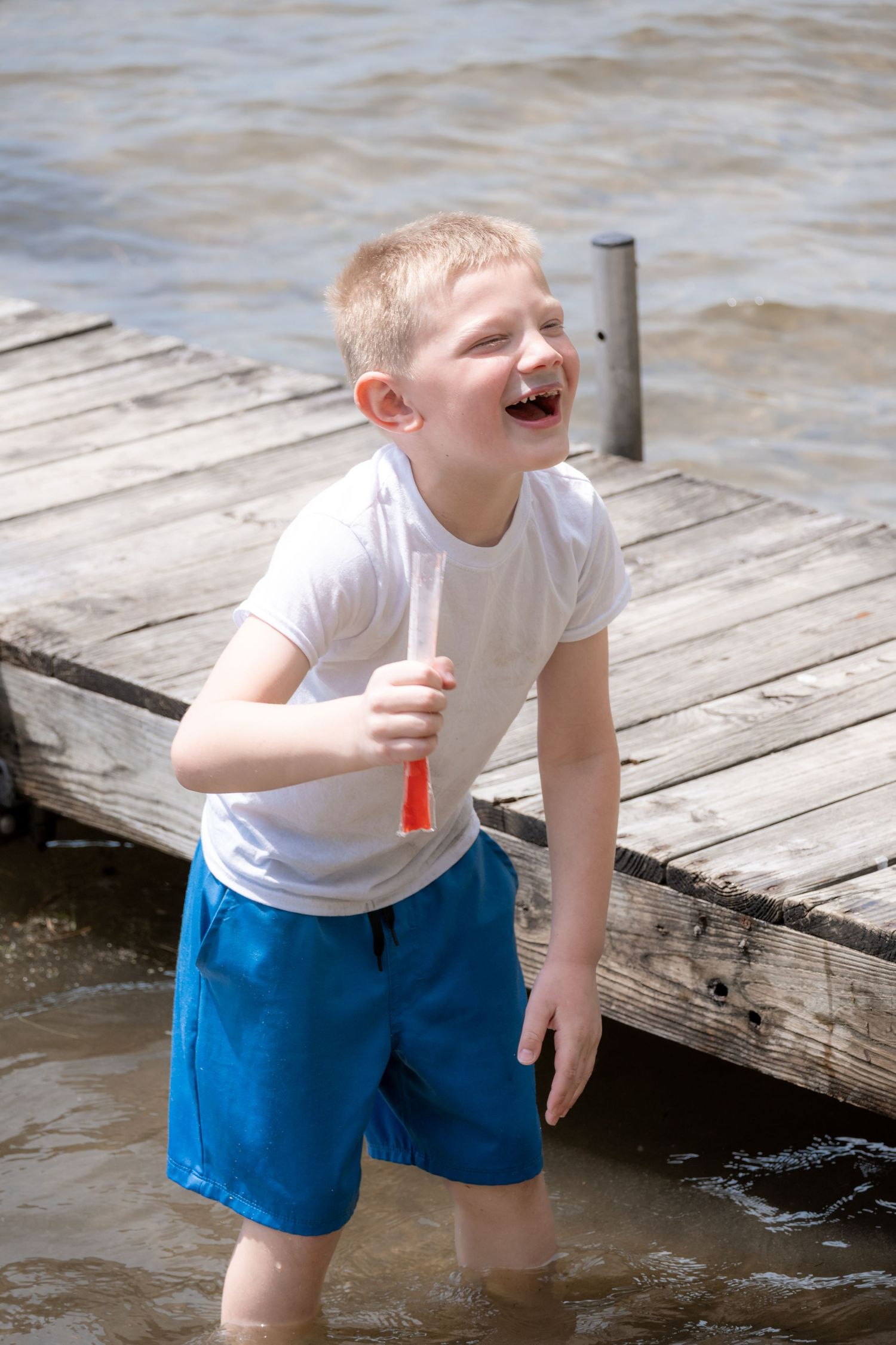 Laughing young boy in white t-shirt and blue shorts standing in water near a wooden dock.