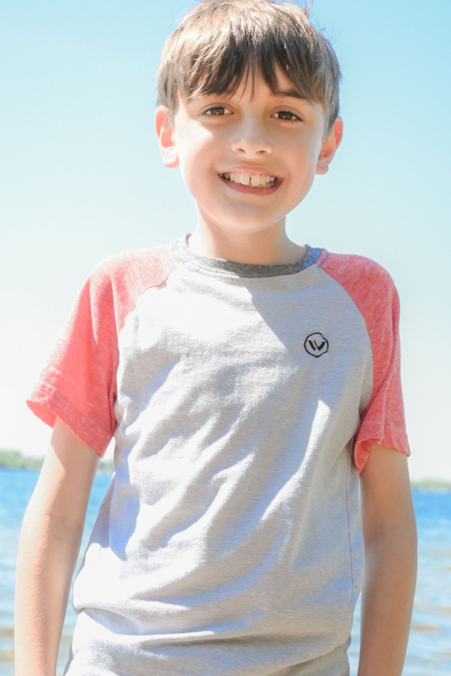 Smiling young boy in a gray and red raglan t-shirt standing outdoors near a lake on a sunny day.