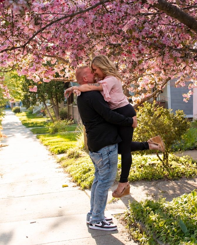 Man lifting woman for a kiss under blooming pink cherry blossom tree on a sunny sidewalk.