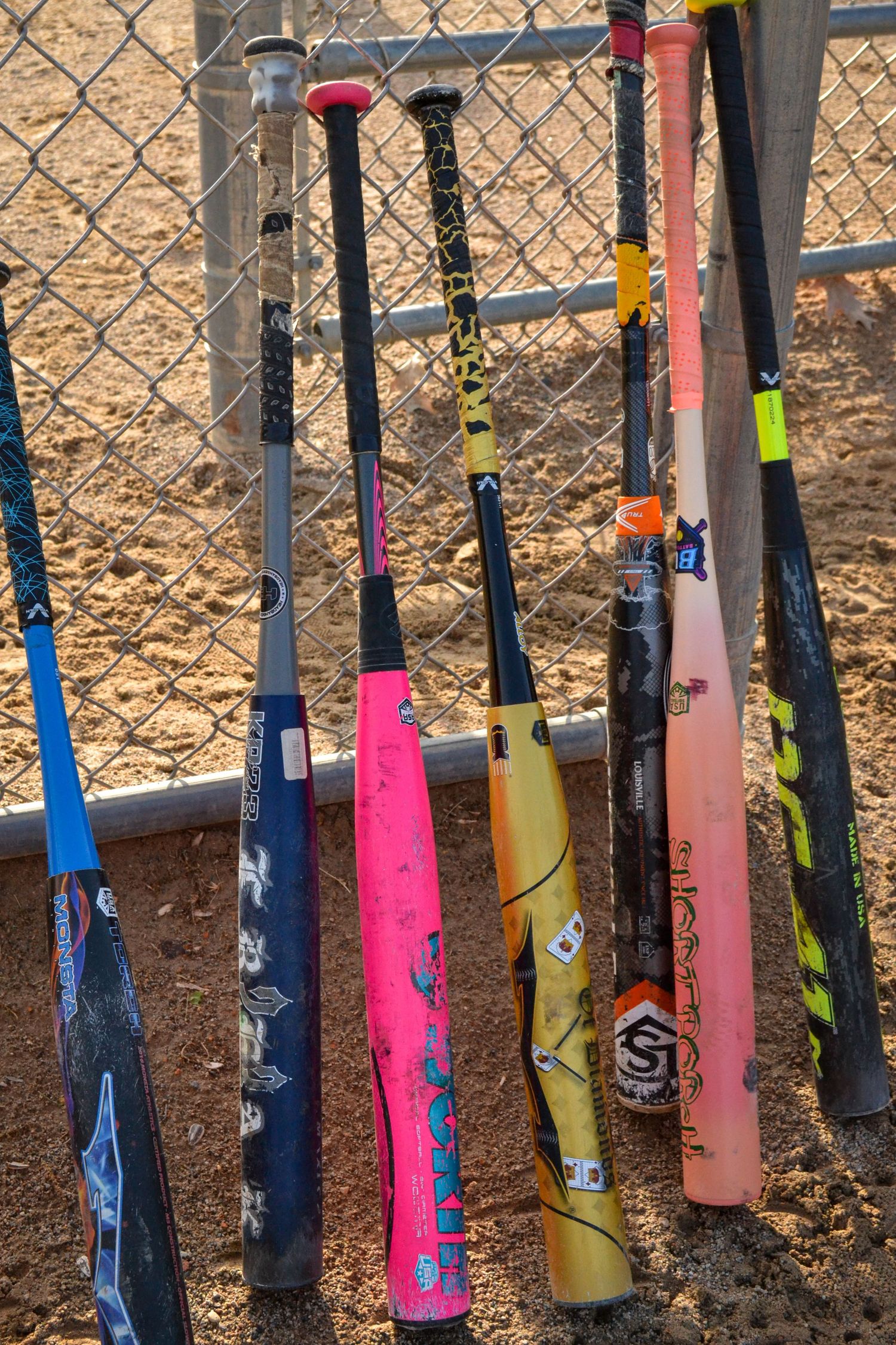 Colorful collection of softball bats leaning against a chain-link fence on a sandy baseball field.