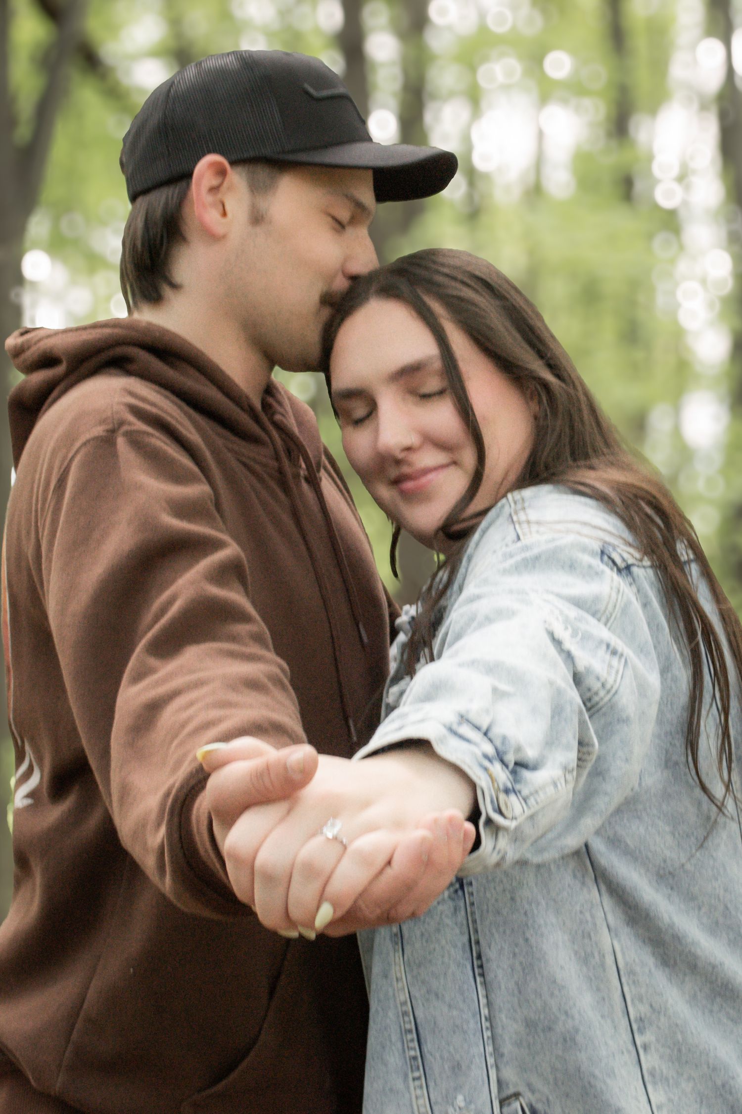 A couple embraces tenderly in a forest, the woman wearing a diamond engagement ring.
