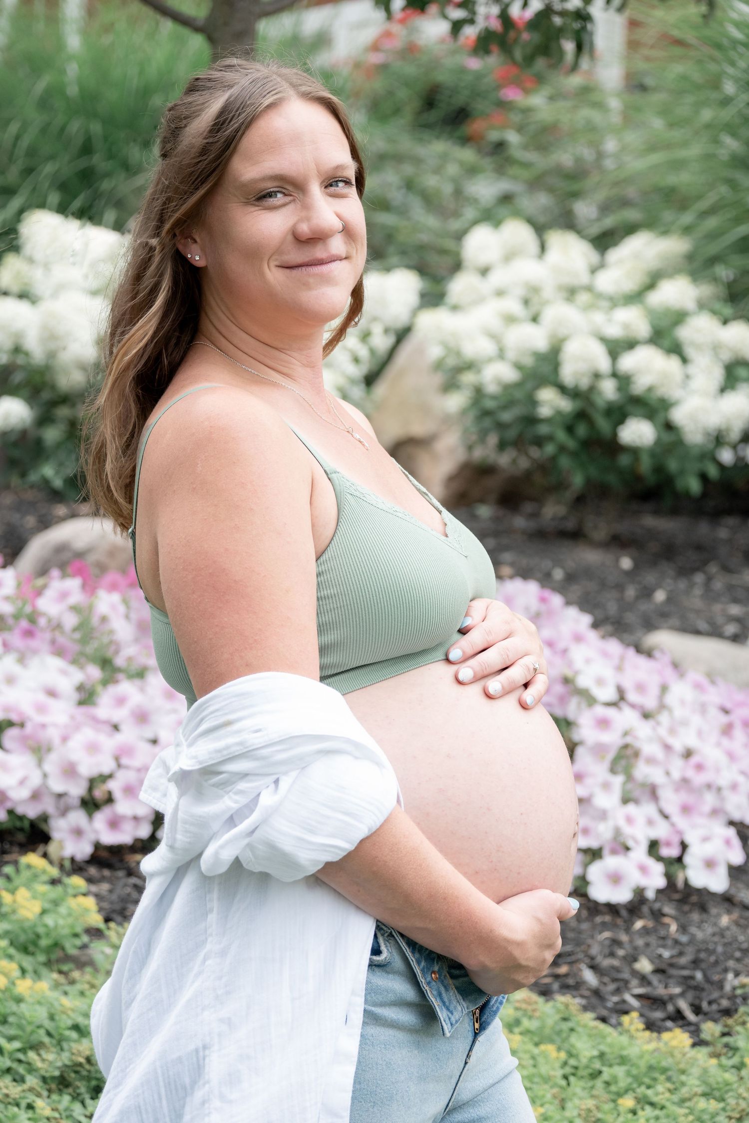 Pregnant woman in sage green bralette and jeans cradling her baby bump outdoors among blooming flowers.