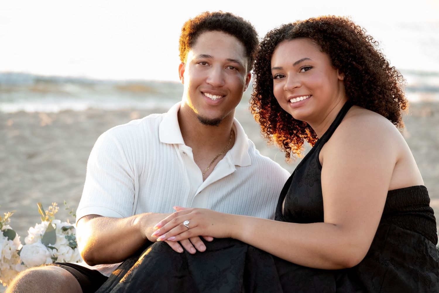 Happy couple sitting together on a sandy beach, smiling and holding hands in warm golden sunlight.