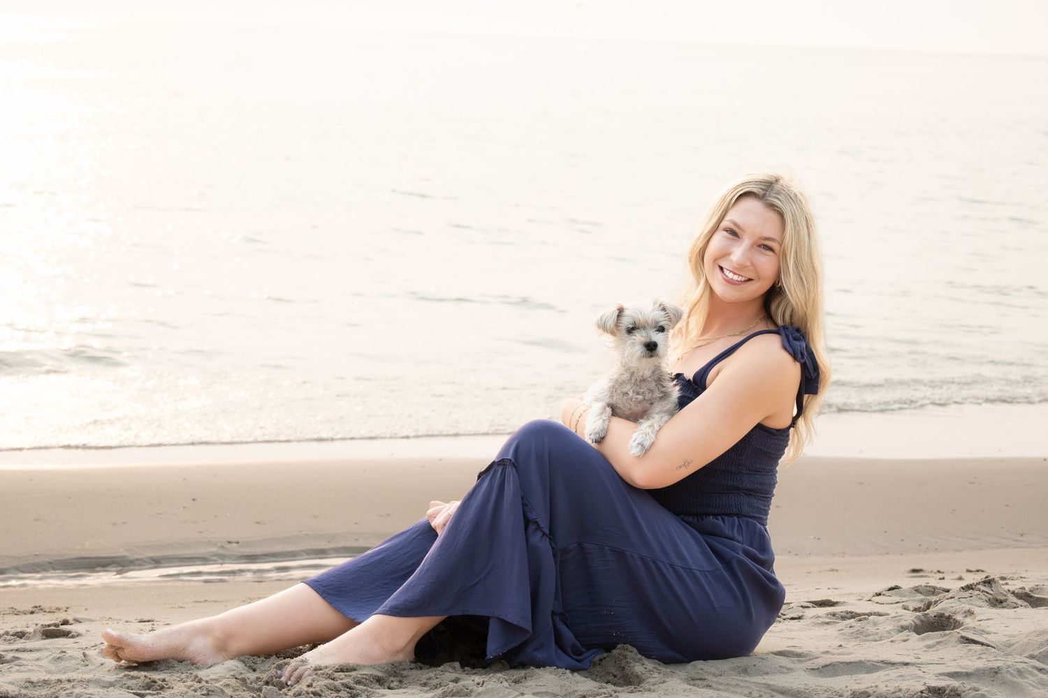 Smiling blonde woman in navy dress sitting on beach sand holding a small schnauzer dog near ocean waves.